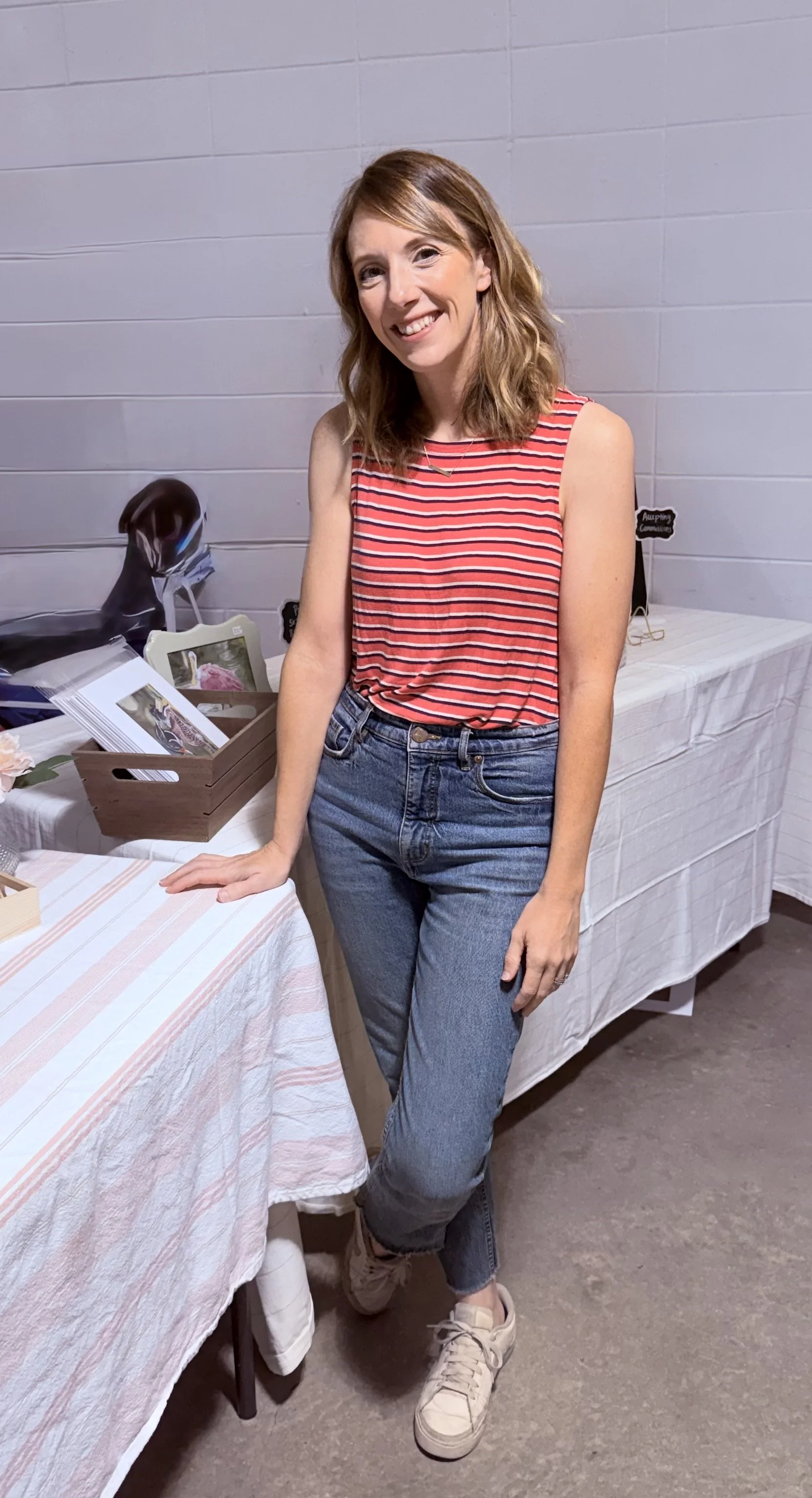 A woman with shoulder-length red hair wearing a red and white striped sleeveless top and blue jeans standing next to a table with various items on it, smiling at the camera.