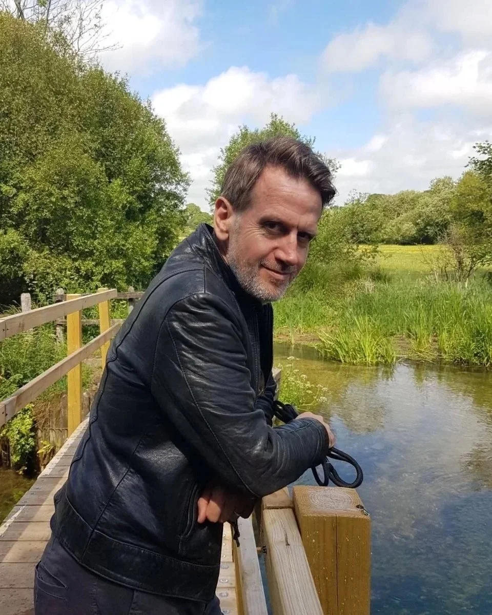 A man in a black leather jacket leaning on a wooden railing by a small river in a lush green landscape.
