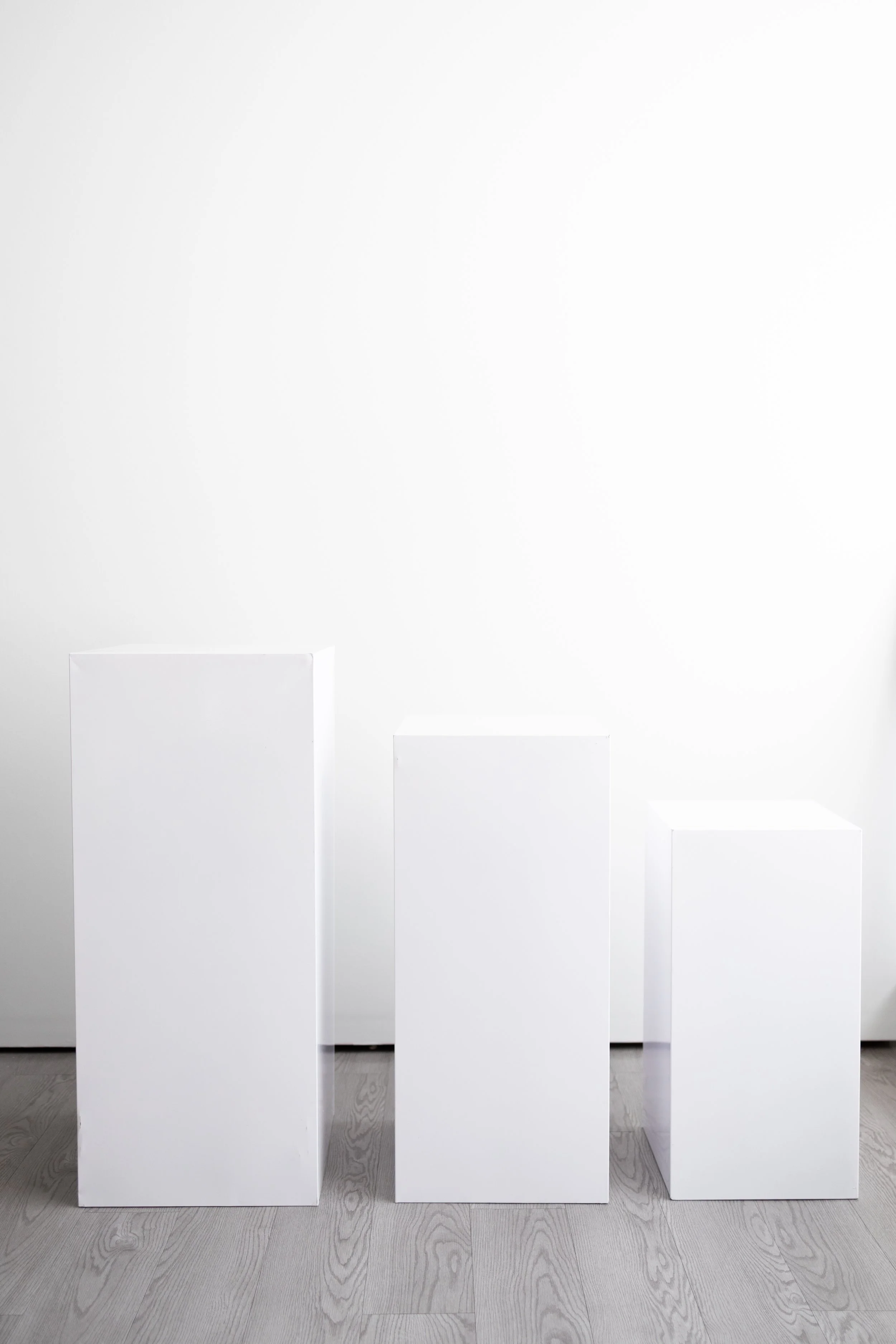 Three white display pedestals of varying heights are arranged in front of a plain white background in a minimalistic setting.