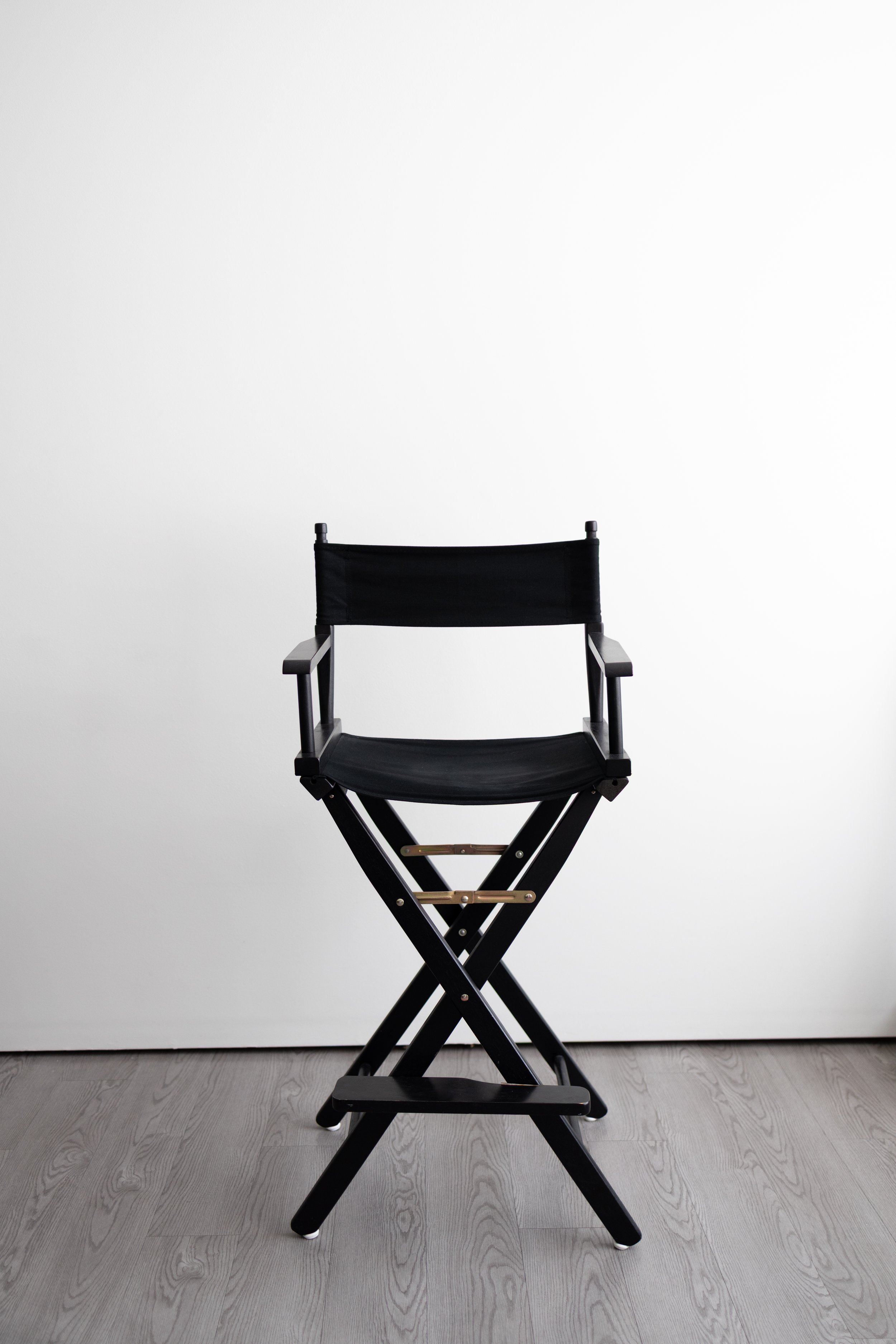 A black director's chair with a wooden frame, positioned in front of a plain white background on a gray wooden floor.