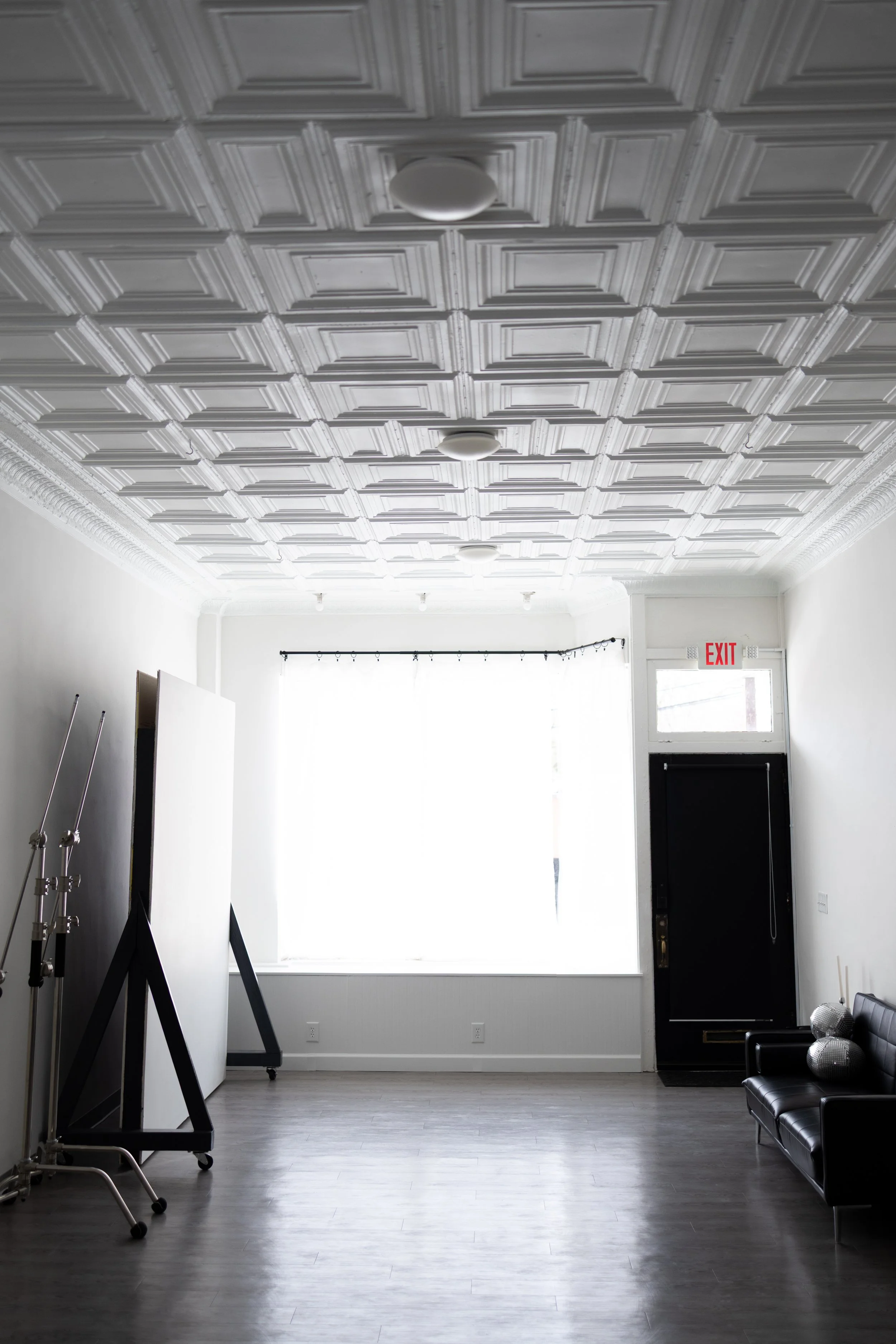 Interior of a room with a decorative ceiling, black door, large window with sheer curtains, black leather sofa, and photography equipment including lights and a diffuser.