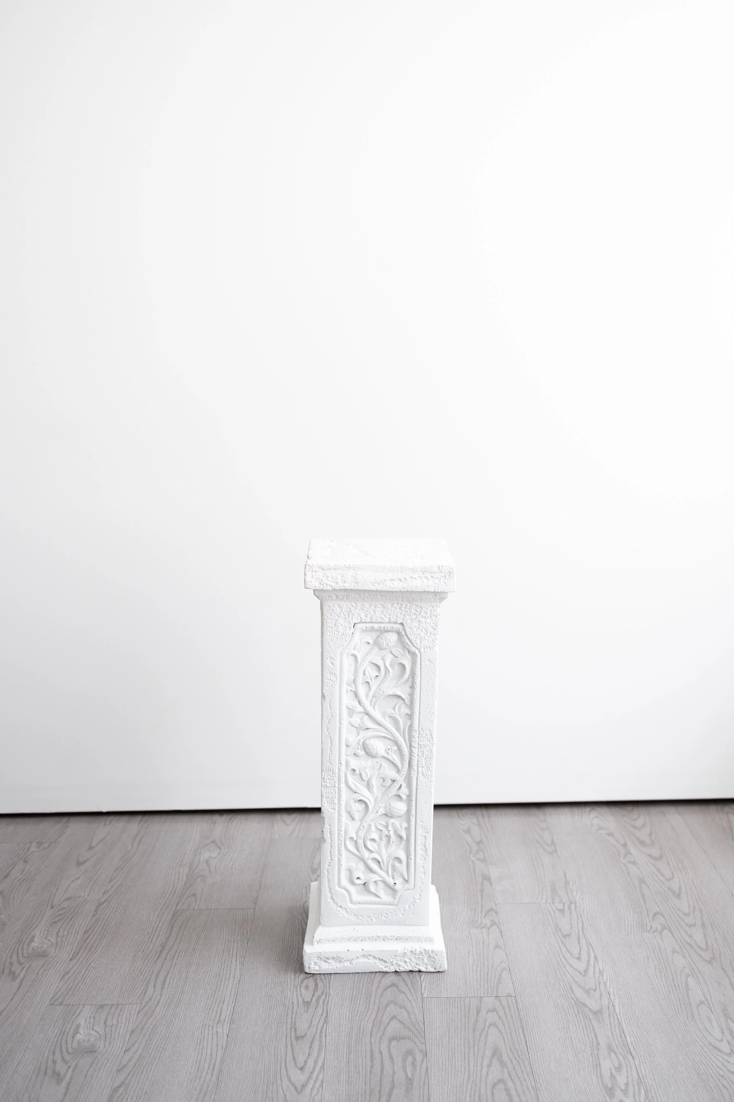 White decorative pedestal with intricate designs, positioned on a gray wooden floor against a white wall.