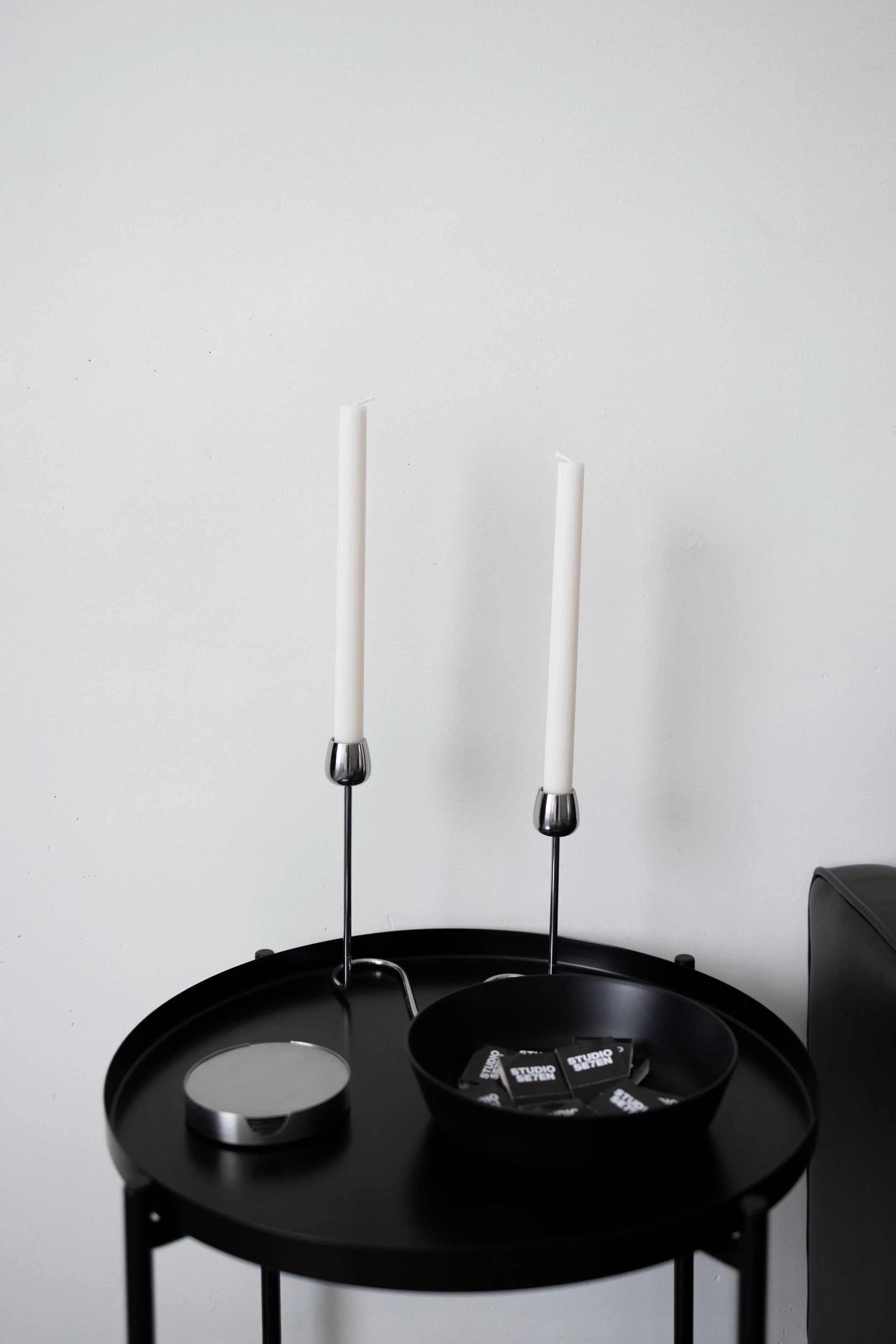 A black side table with white candles on silver candle holders, a small round mirror, and a black bowl containing black and white cards, set against a plain white wall.