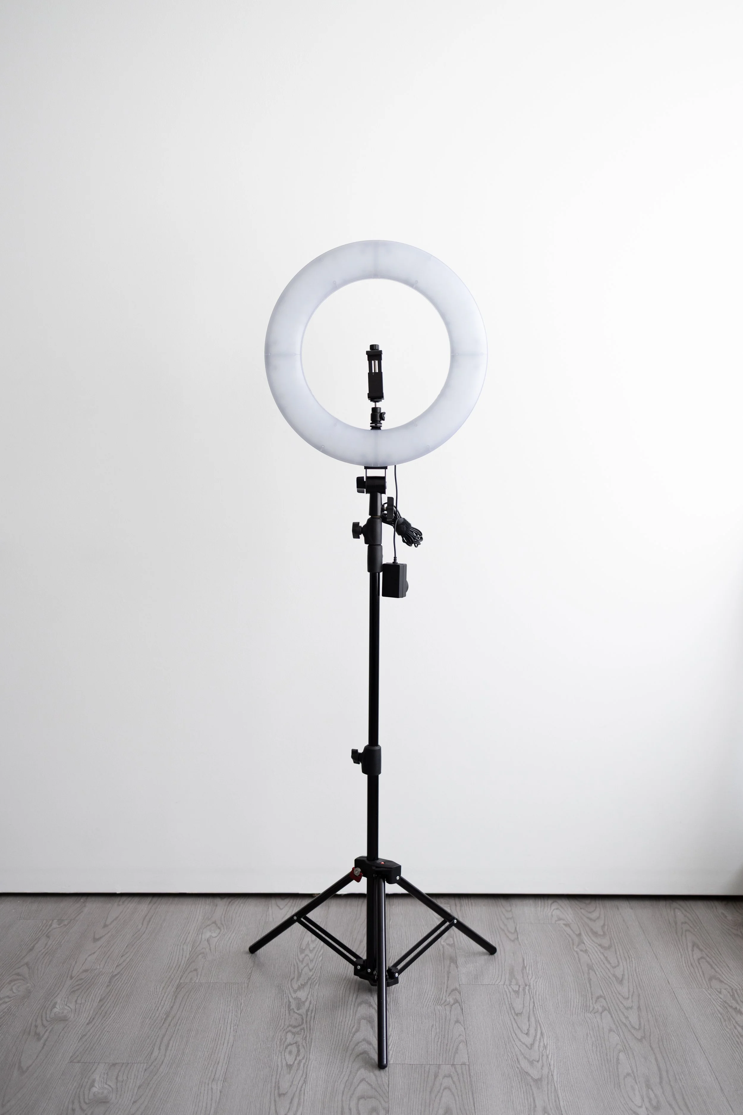 A photography ring light on a tripod stand against a white wall.