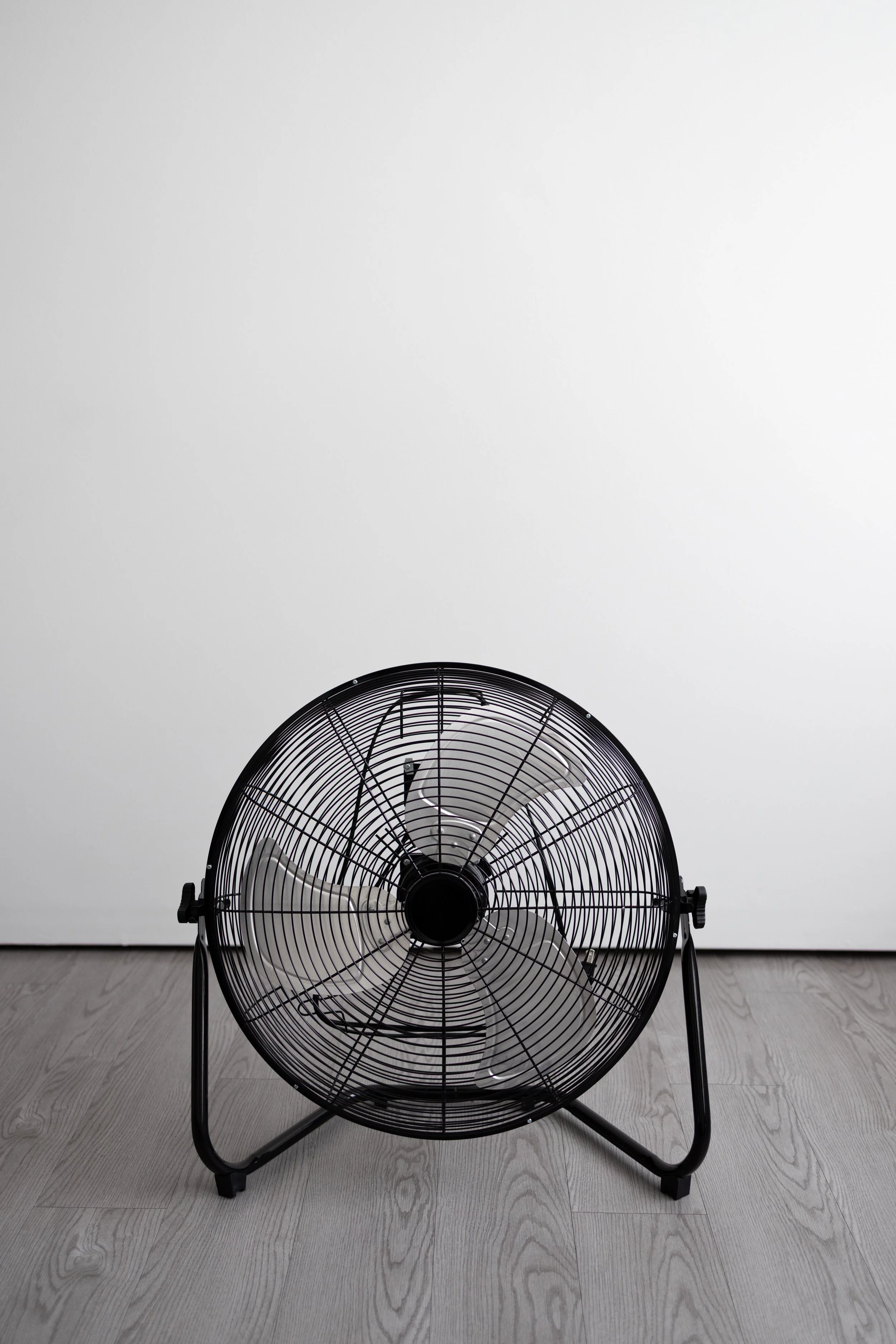 Black floor fan in front of a plain white wall on a gray wooden floor.
