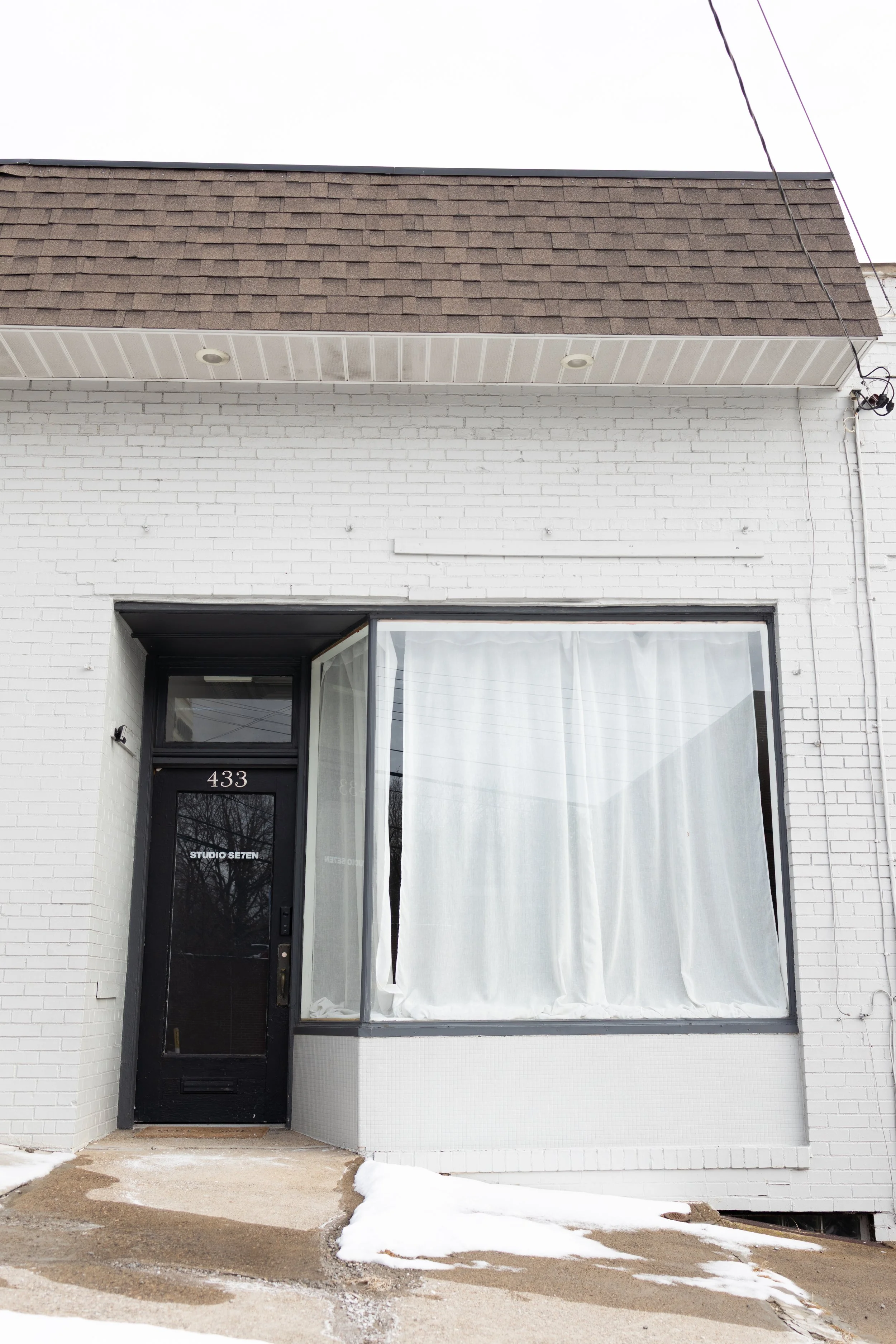 A storefront with a black door labeled 'Studio Seven' at 433, white brick walls, a large window with white curtains, and a snow-covered sidewalk.