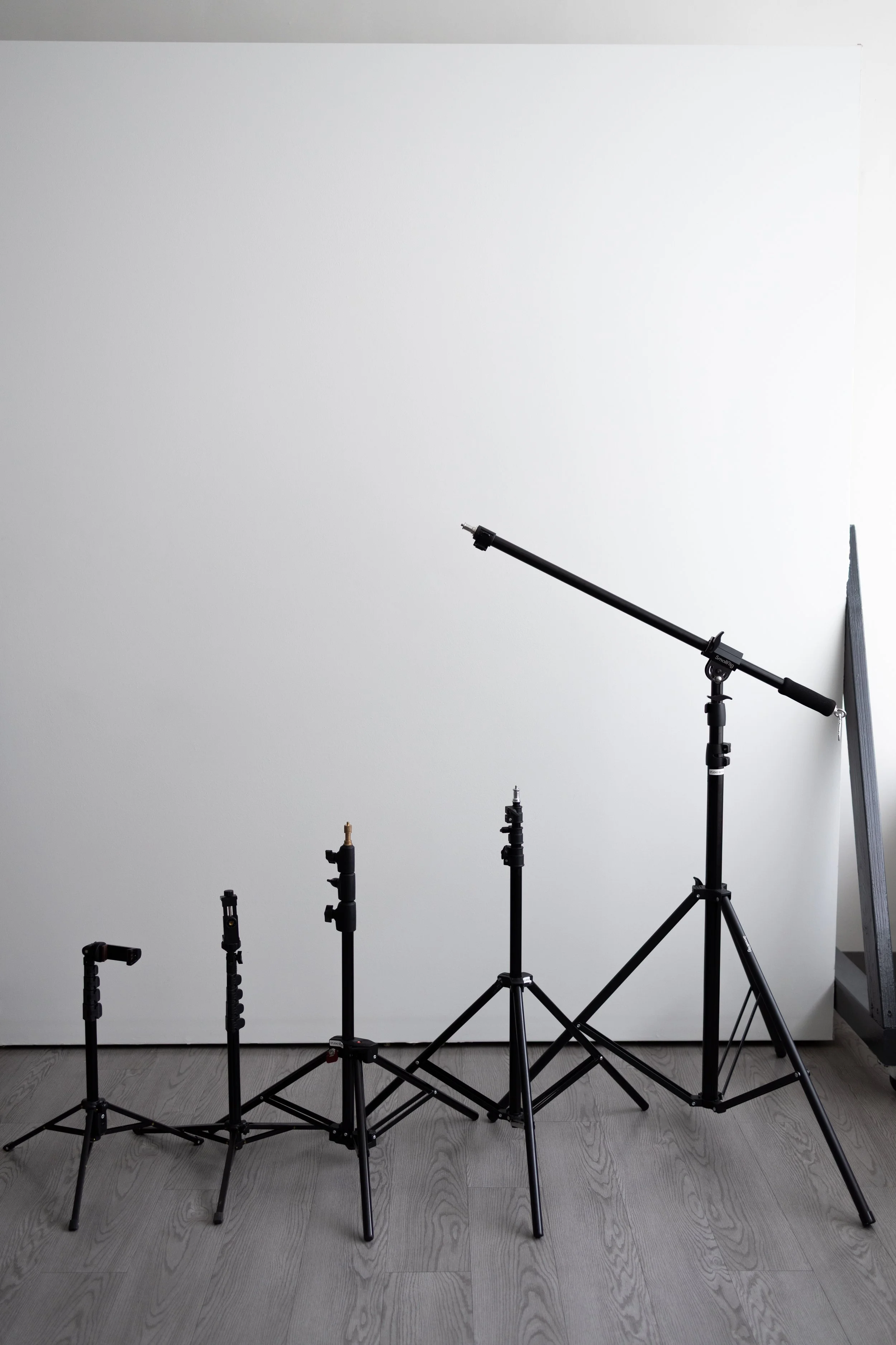 Five studio light stands of varying heights arranged in front of a plain white wall on a wooden floor.