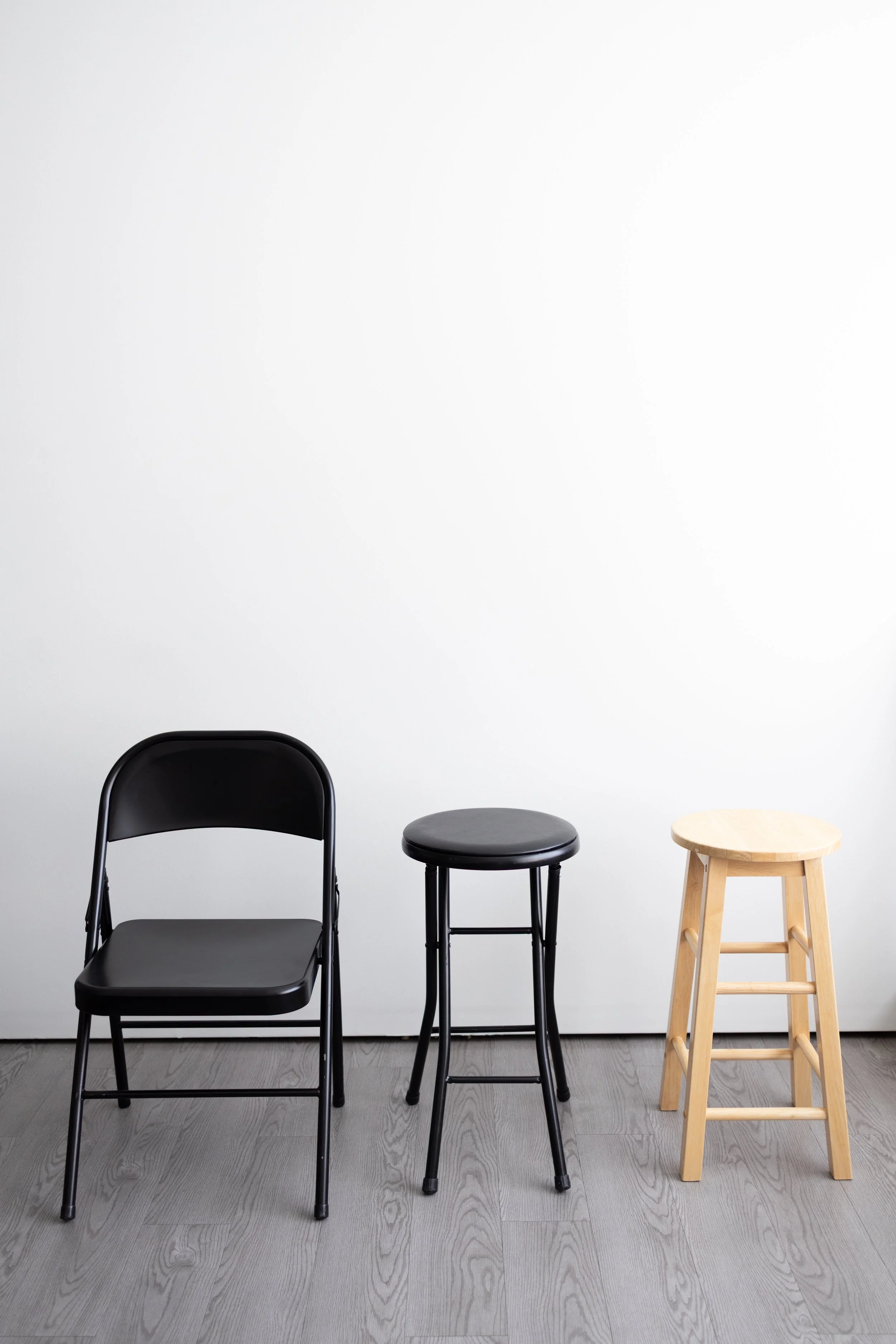 Three seats of different types placed in a row against a plain white wall and a light grey wooden floor: a black folding chair, a black round stool, and a light wooden barstool.