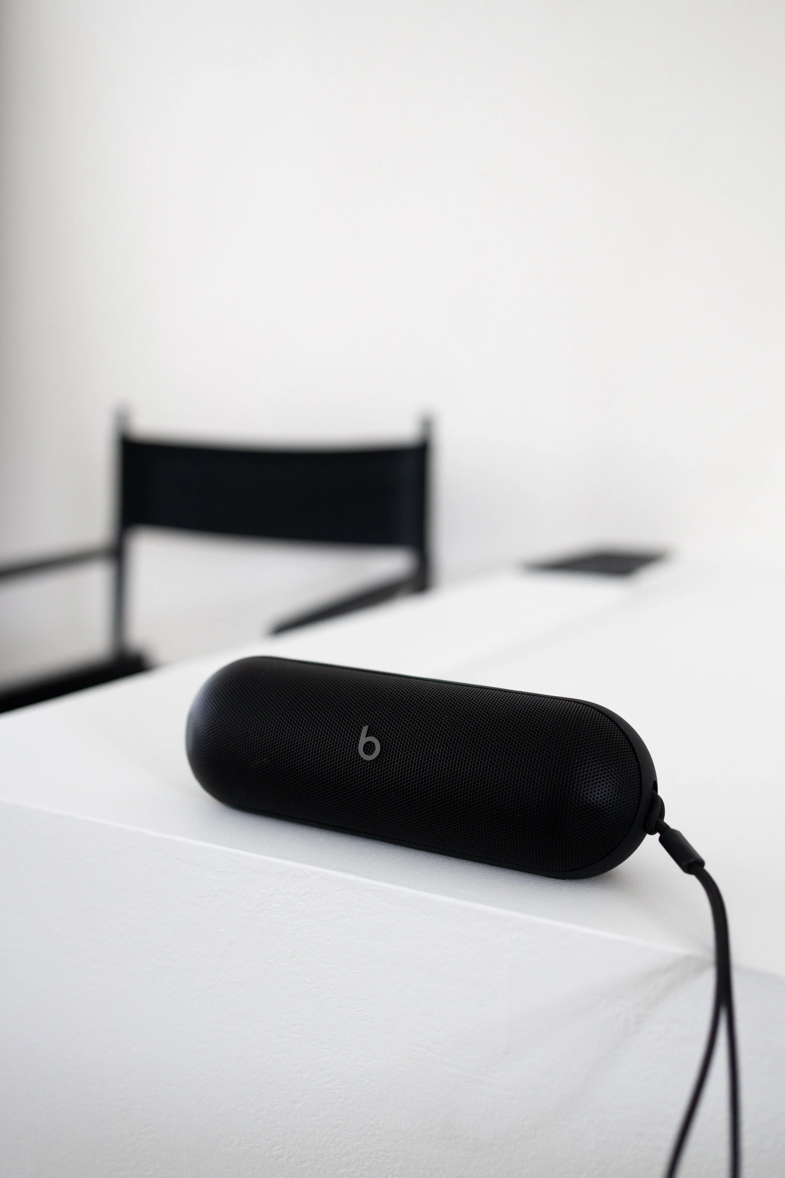 Black portable speaker with a 'b' logo on a white table, with a black chair and a white wall in the background.