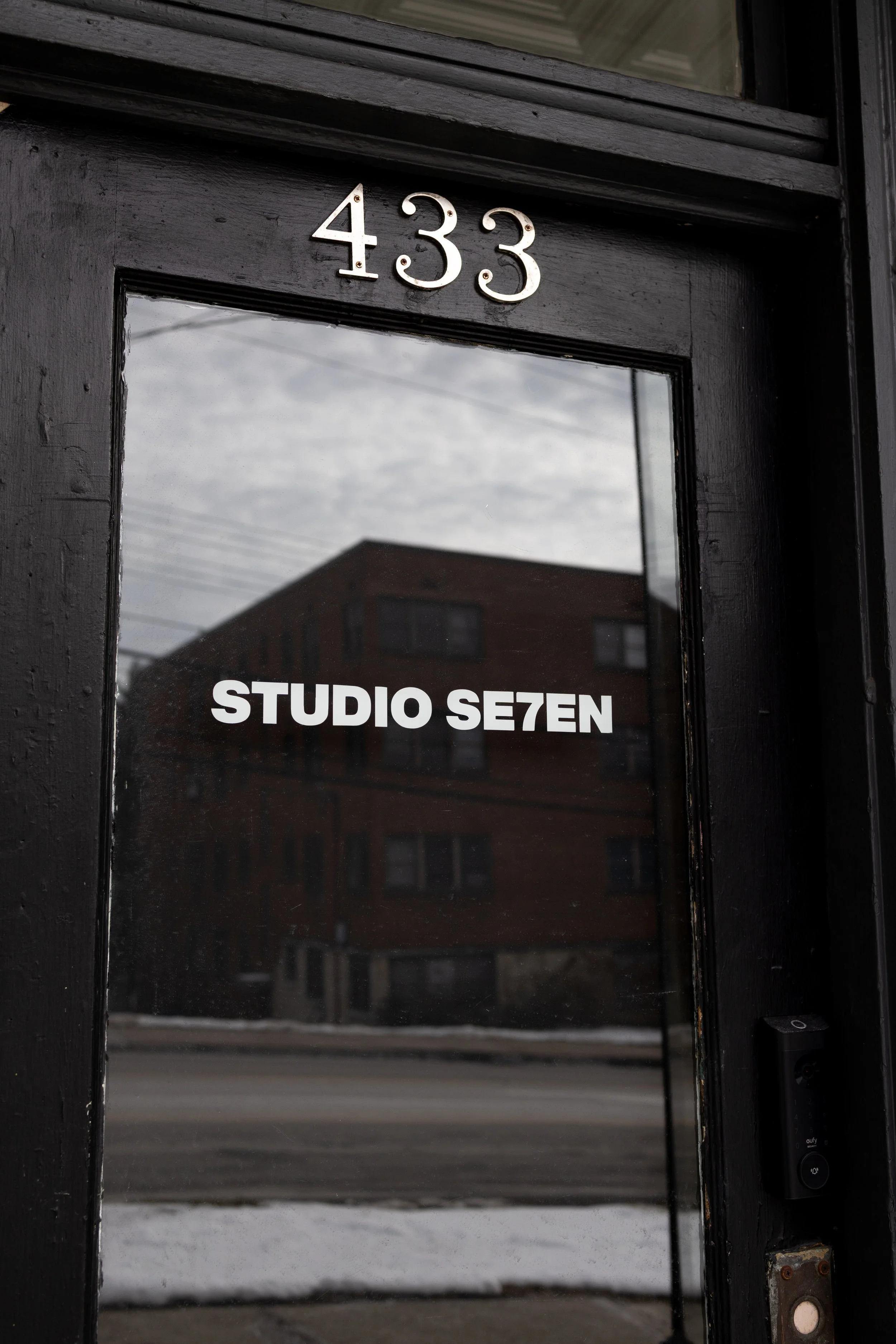 Black door with house number 433 and a glass window reflecting the street and a building, with white sign reading "STUDIO SE7EN".