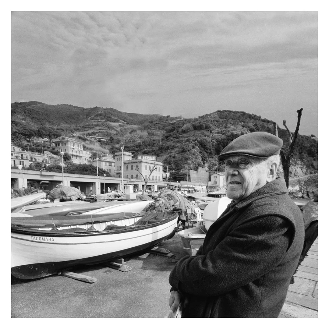 A black and white photo of an elderly man wearing a cap and coat glancing sideways near boats on a waterfront with houses on hills in the background.