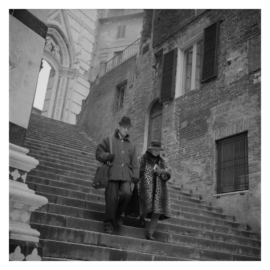 Two people walking down a set of outdoor stone stairs in an old European city with brick buildings and arched windows.