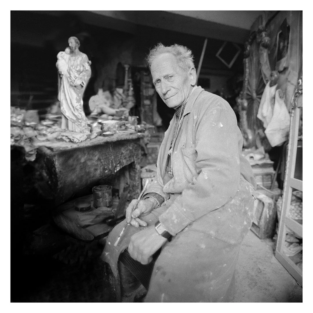 An elderly man with a work apron sitting in a cluttered workshop, surrounded by sculptures and art supplies.