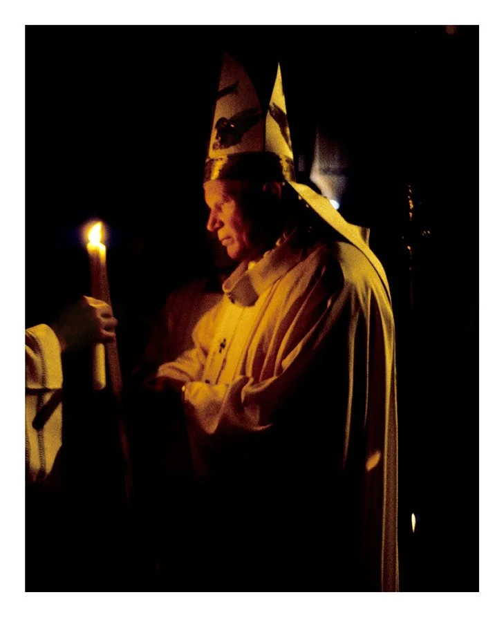 A person dressed in religious robes and a tall, pointed hat, holding a lit candle, appears to be engaged in a religious or ceremonial activity in a dark setting.