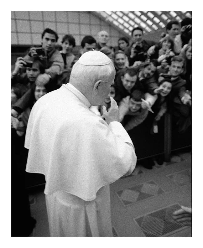 Pope Francis praying with hands clasped in front of a crowd of spectators behind a barrier.