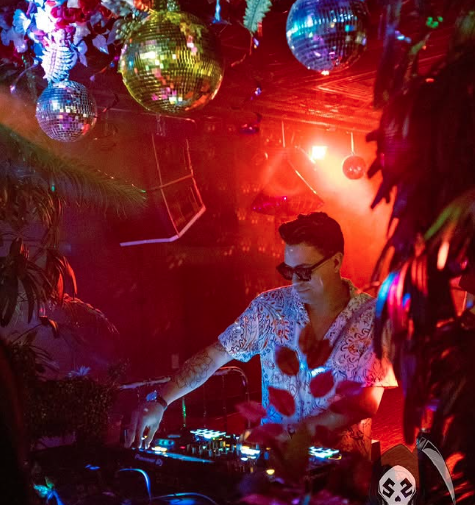 Braulio, aka DJ Bayo, performing at a dimly lit nightclub with colorful lights, disco balls, and tropical decorations.