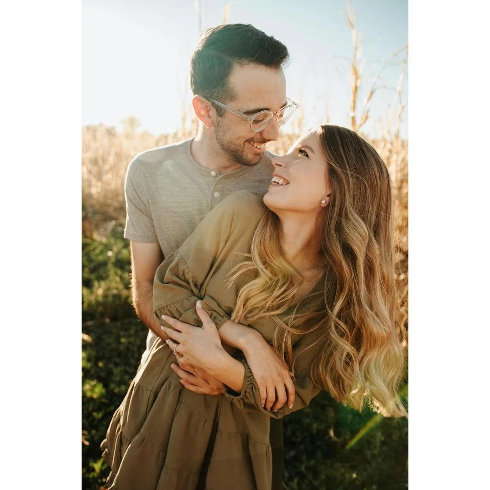 A couple embracing and smiling at each other in a sunny outdoor setting with tall grass in the background.