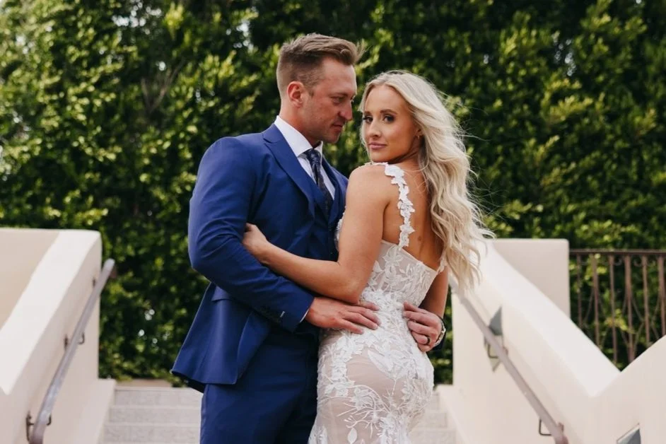 A man in a blue suit and a woman in a white wedding dress embracing on a staircase with green foliage in the background at Secret Gardens by Wedgewood Weddings in Arizona