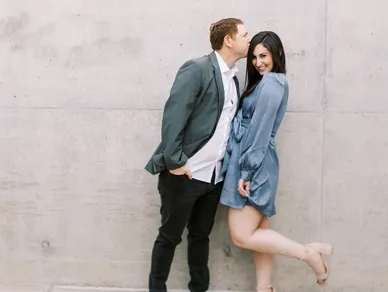 A couple posing against a concrete wall, with the man wearing a blazer and kissing the woman's forehead, and the woman wearing a blue dress, smiling.