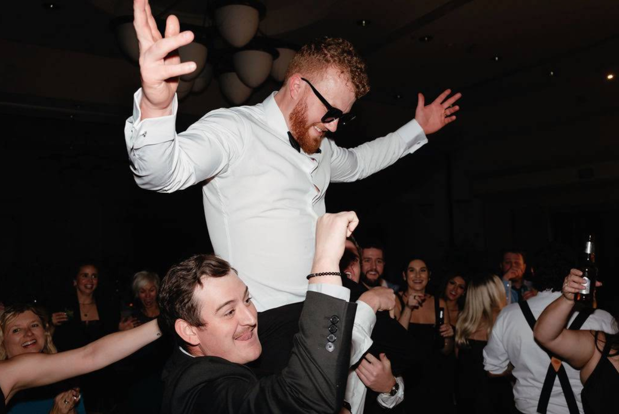 People celebrating at a wedding, with a groom being lifted on a friend's shoulders. The crowd is smiling and holding drinks.
