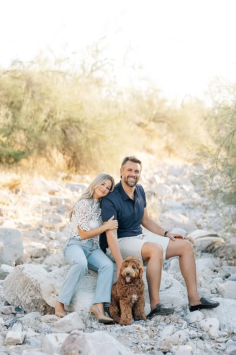 A couple sitting on a rock in a natural outdoor setting with a brown fluffy dog at their feet, surrounded by dry vegetation and rocks.
