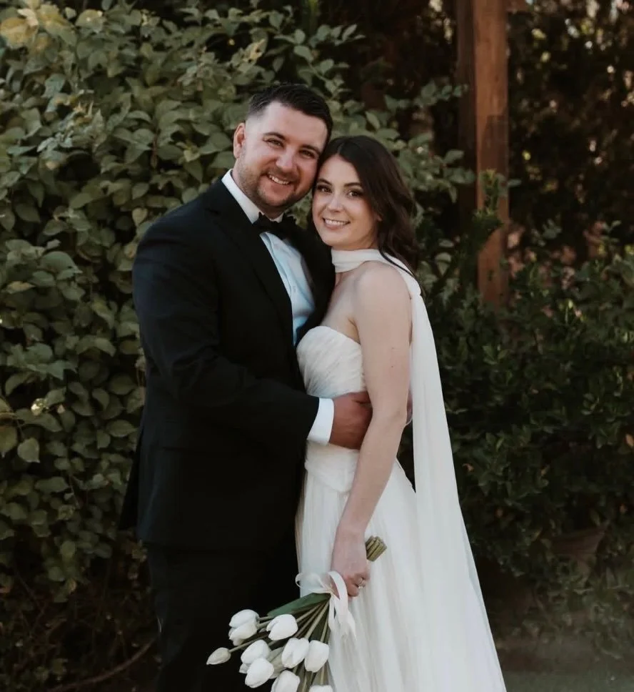 A couple sitting on a kitchen counter, smiling at each other. The man is wearing a blue shirt and jeans, while the woman is in a black top, denim shorts, a brown cardigan, and a black hat. The kitchen has white cabinets, a stainless steel stove, and decorative items like a plant and cutting boards.