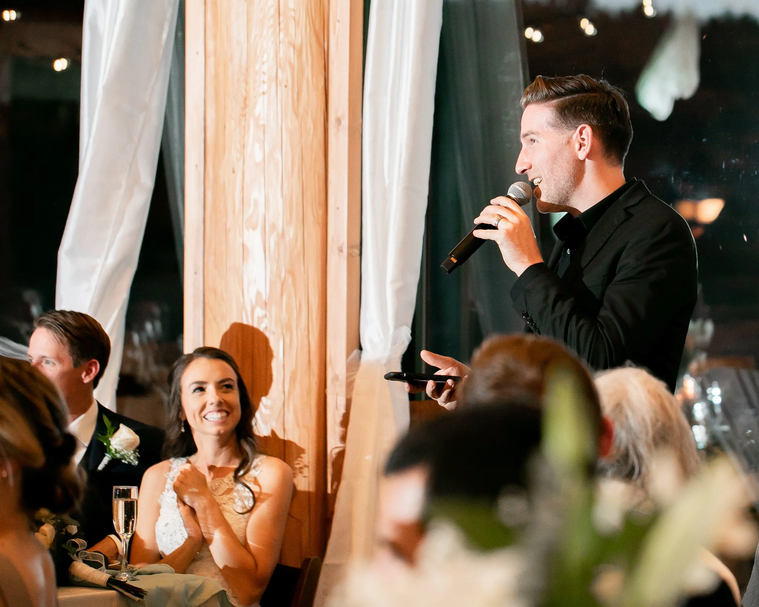Wedding reception with a smiling bride and a man making a speech using a microphone. The groom and guests are seated nearby, and the venue is warmly lit with draped curtains and wooden decor.