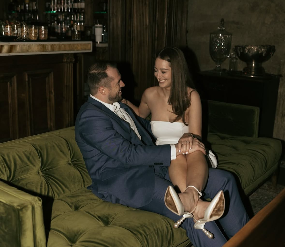 A man and woman sitting closely on a green velvet couch in a dimly lit room, dressed in formal attire, with the woman smiling and resting her legs on the man's lap.