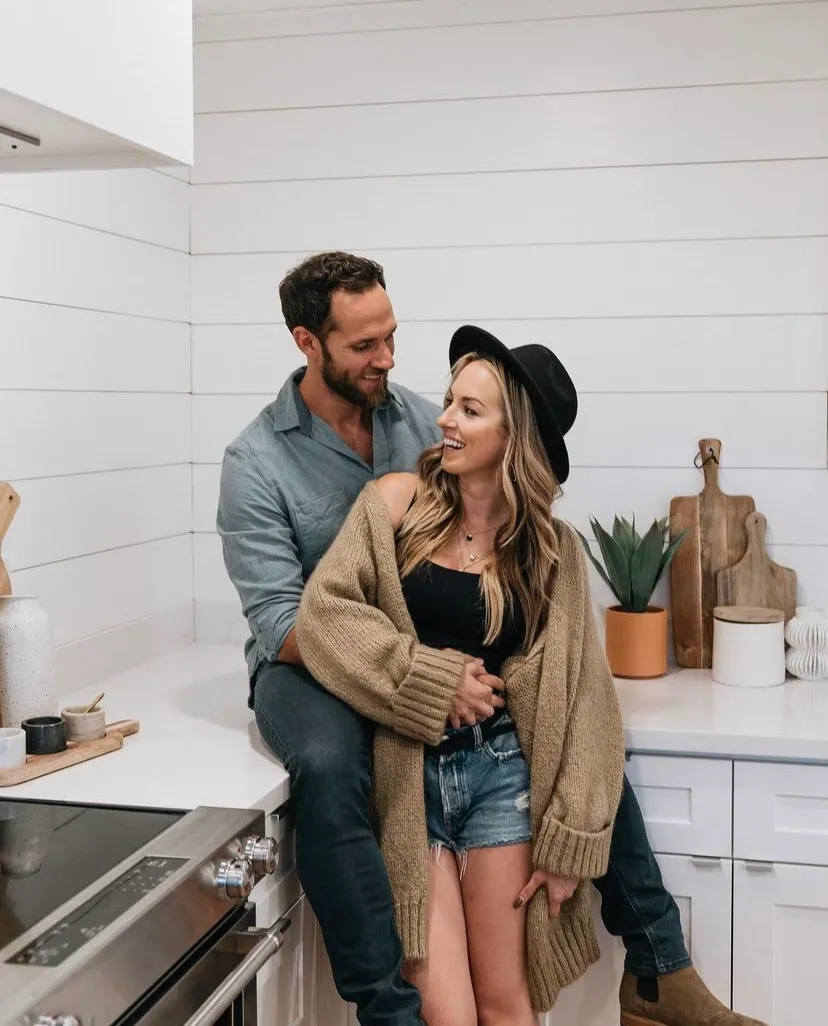 A couple sitting on a kitchen counter, smiling at each other. The man is wearing a blue shirt and jeans, while the woman is in a black top, denim shorts, a brown cardigan, and a black hat. The kitchen has white cabinets, a stainless steel stove, and decorative items like a plant and cutting boards.