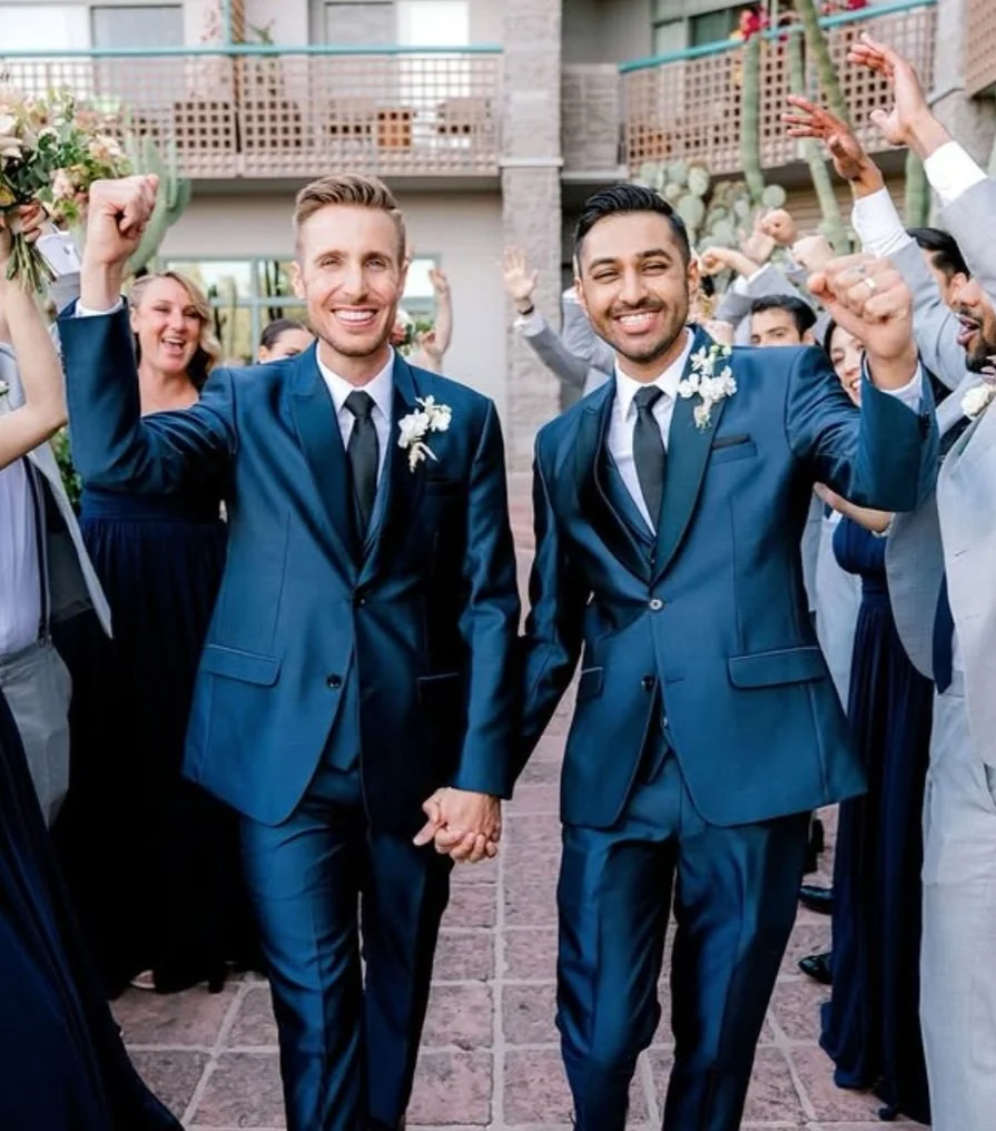Two grooms in blue suits holding hands, celebrating with friends at a wedding at Hyatt Grand Regency in Scottdsale, Arizona