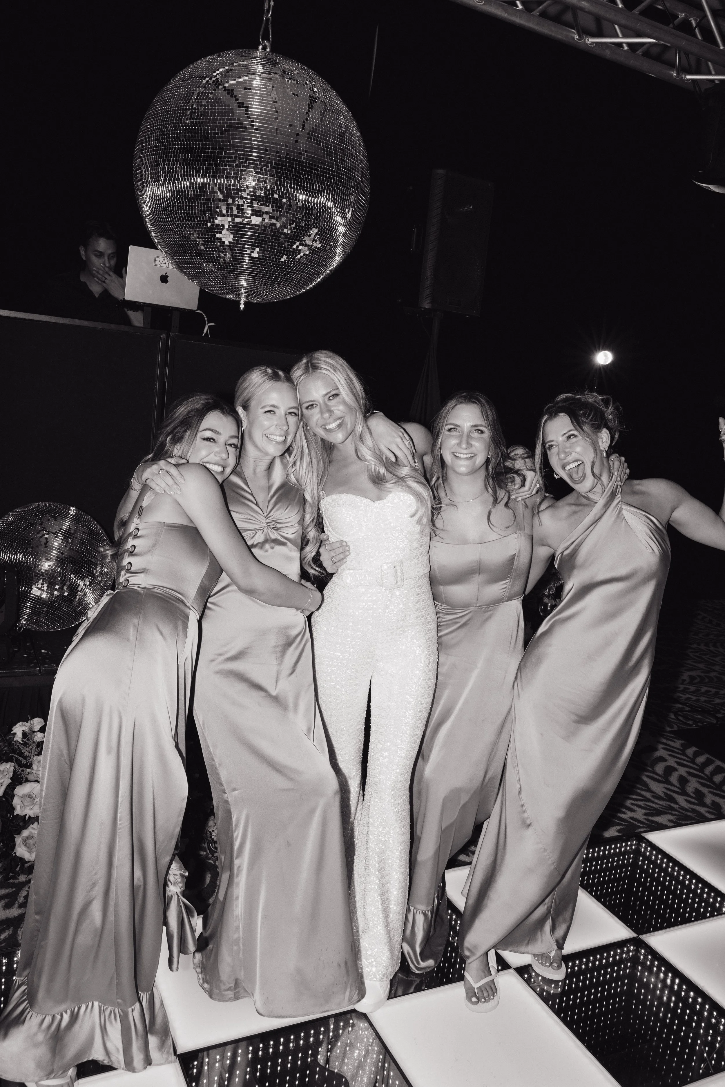 Group of women posing together at an event with a disco ball overhead and a DJ in the background, wearing elegant dresses and a sequined jumpsuit.