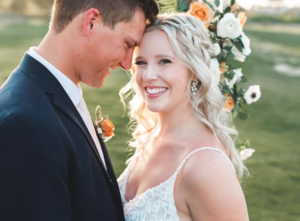 A bride and groom smiling with their heads touching, surrounded by flowers in an outdoor setting at Legacy Golf Club in Arizona