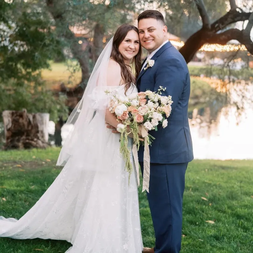 A bride and groom smiling and standing outdoors. The bride is wearing a white gown and holding a bouquet of flowers, while the groom is in a blue suit. They are in a park-like setting with trees and a pond in the background.