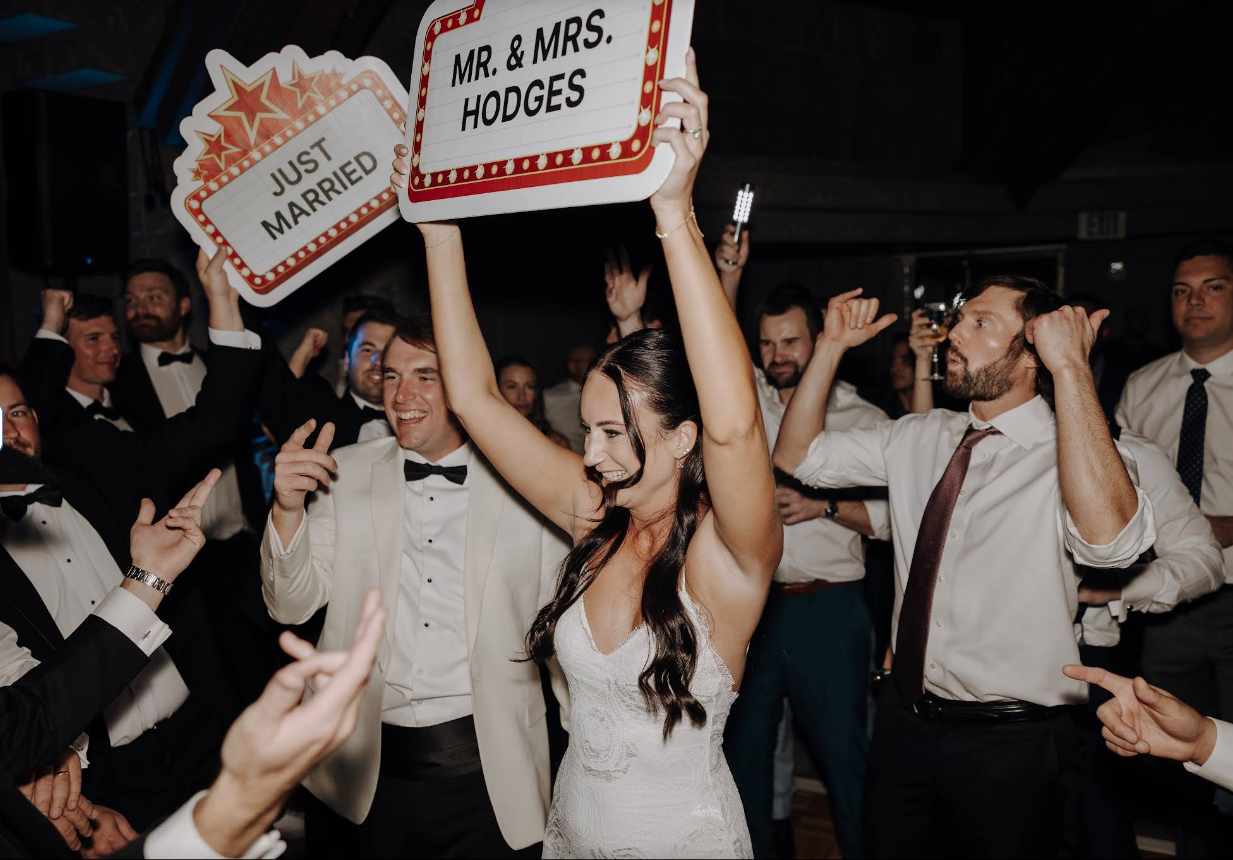 A bride in a wedding gown holds a sign reading 'MR. & MRS. HODGES' while a groom in a white tuxedo smiles next to her.  One guest holding a 'JUST MARRIED' sign. The background shows a lively wedding reception at the Biltmore in Phoenix, Arizona.
