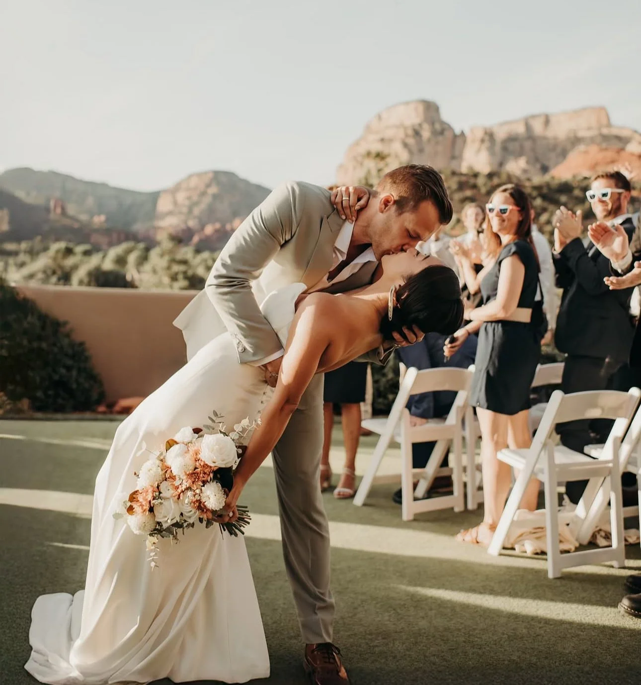 Bride and groom kissing outdoors at a wedding ceremony with guests clapping in the background.