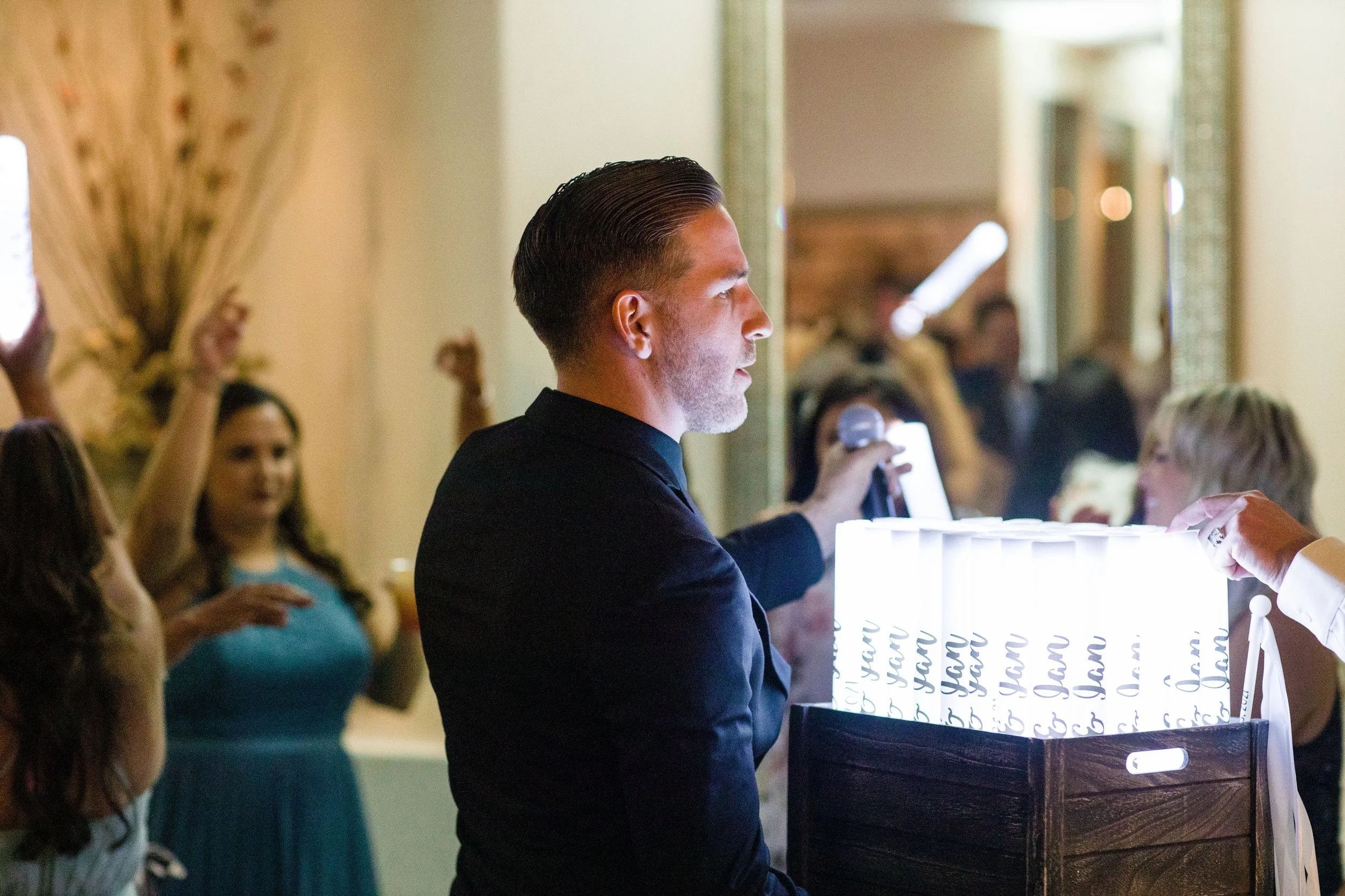 Tyler, aka DJ Munition,  in a suit holding a microphone and a box of light sticks at an event, with people in the background raising their arms.