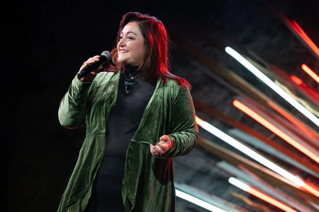Samantha Talora, a woman with brown hair, smiling, wearing a black top and green jacket, holding a microphone, on stage with colorful light streaks in the background.