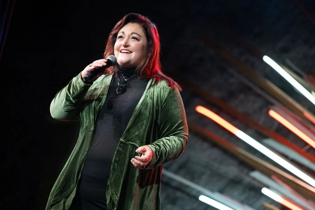A woman with reddish-brown hair singing into a microphone on stage, wearing a green velvet blazer and a black outfit, with colorful stage lights in the background.