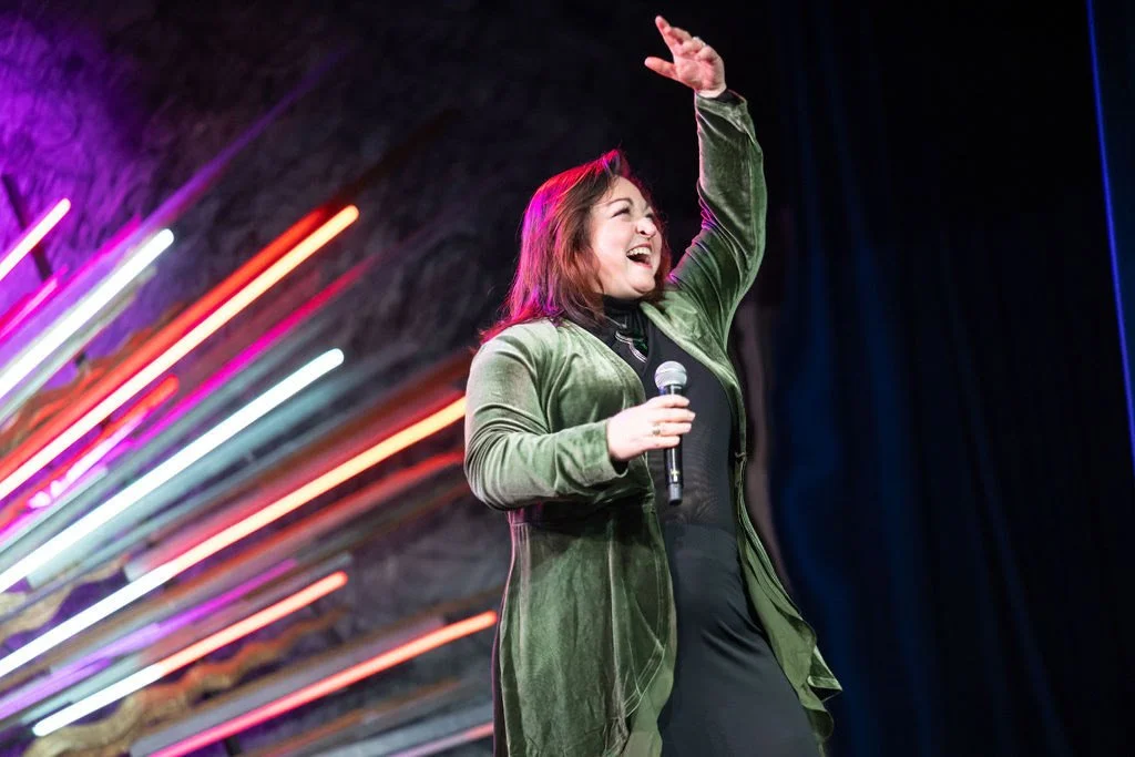 A woman smiling and laughing while holding a microphone on stage, with colorful neon lights in the background.