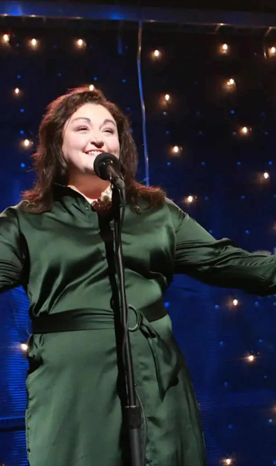 A woman with brown hair singing into a microphone on stage with a starry background.