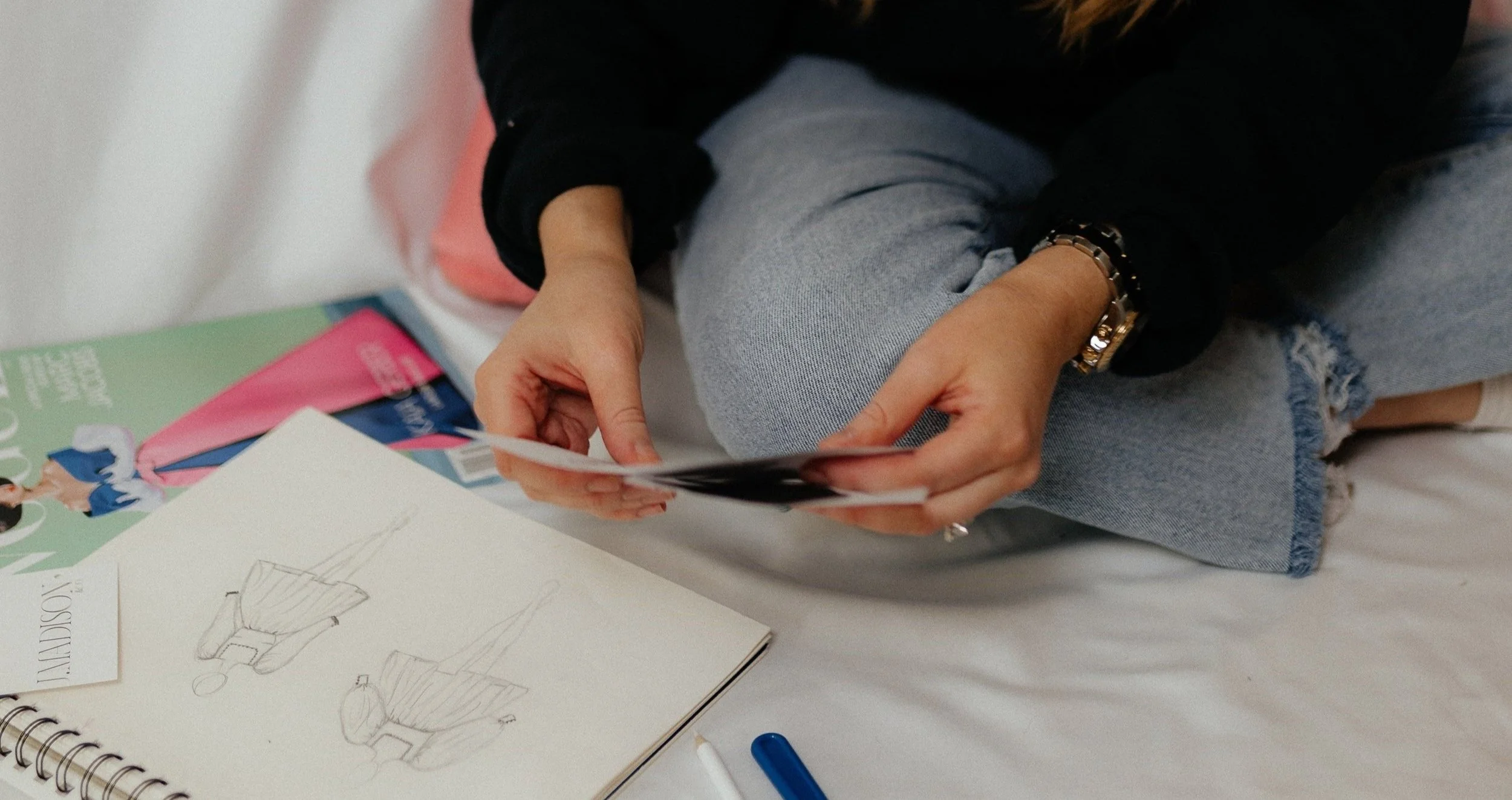 Person holding a photo, sketchbook with fashion drawings, Vogue magazine, and a pen.