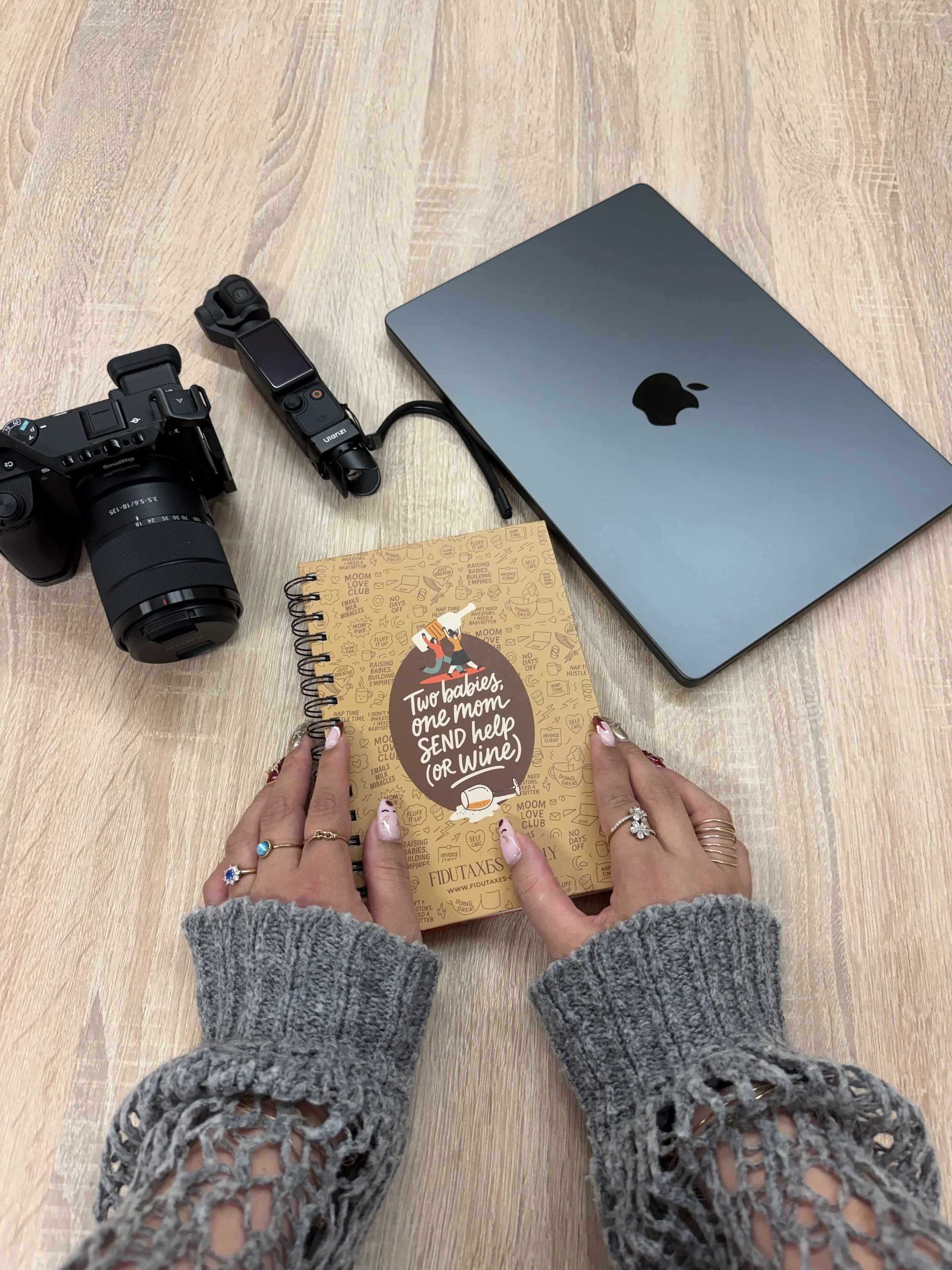 Mains tenant un carnet avec la phrase "Two babies, one mom SEND help (or wine)" sur une table en bois clair, avec un ordinateur portable, un appareil photo sony et l'osmo pocket 3