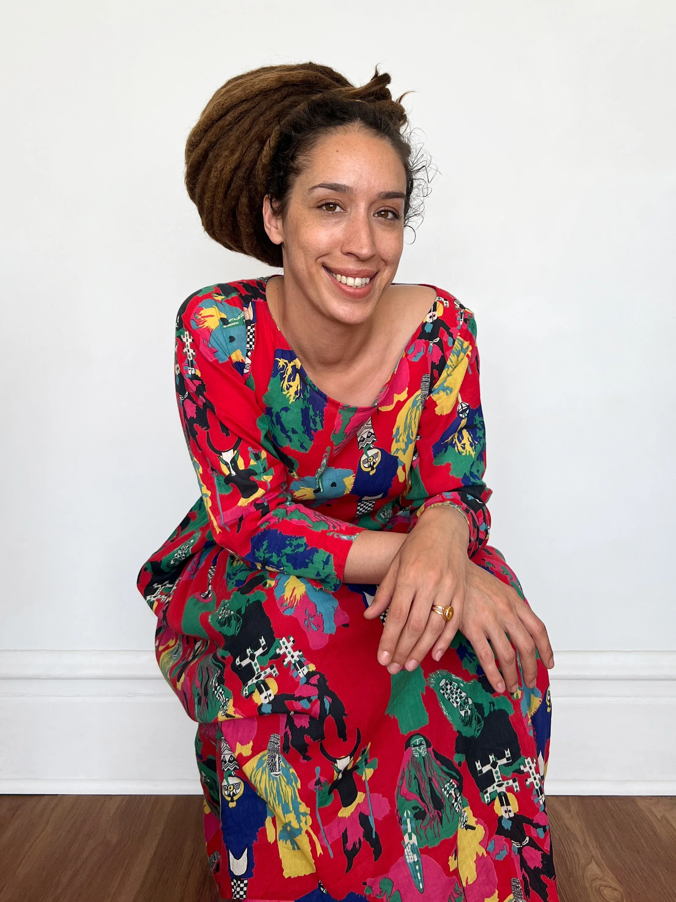 A woman with medium skin tone and dreadlocks, wearing a colorful dress with various abstract and graphic patterns, is smiling and sitting against a plain white wall with a wooden floor.