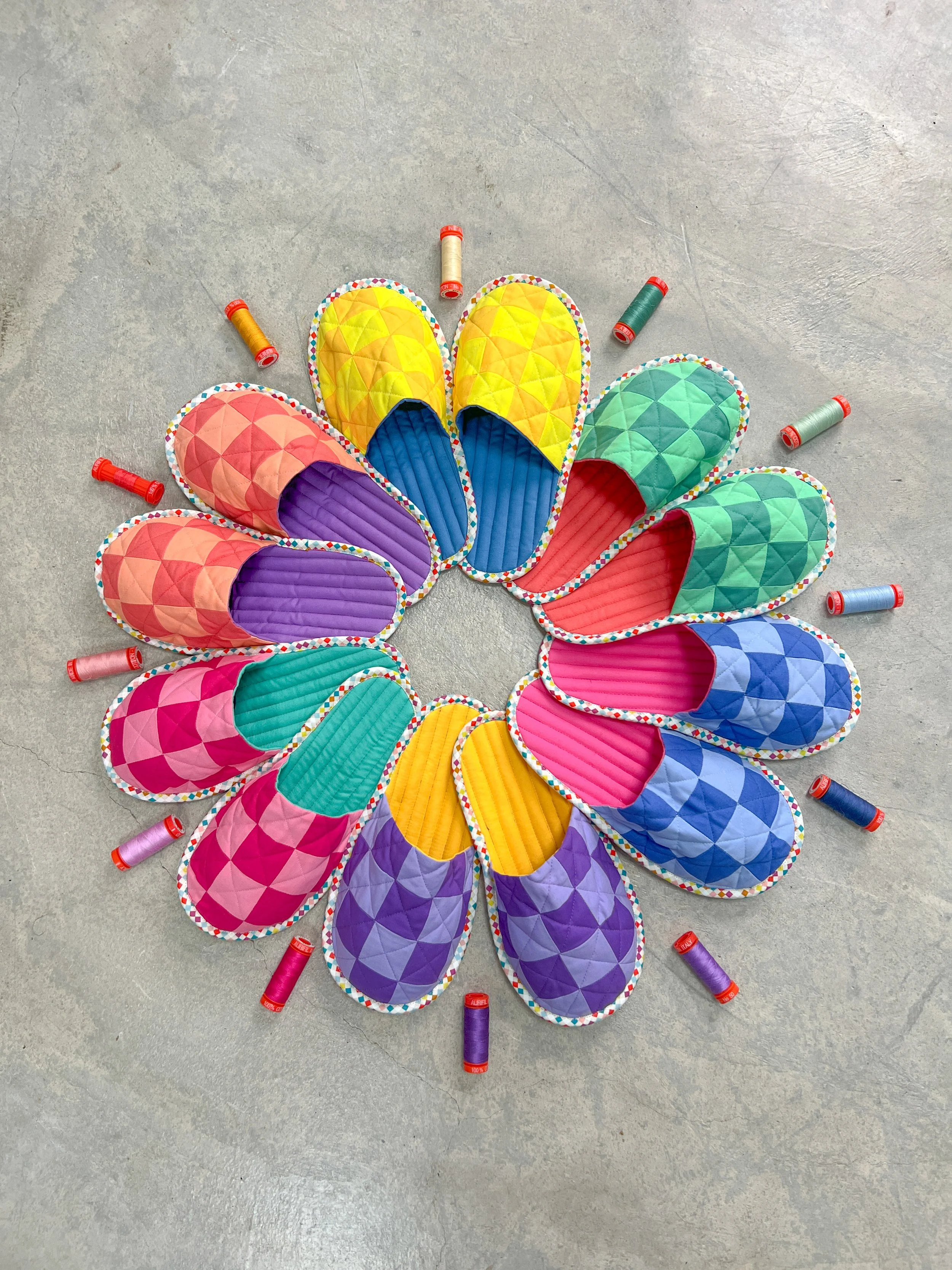 Colorful quilted slippers arranged in a circle on a gray floor, with spools of thread around them.