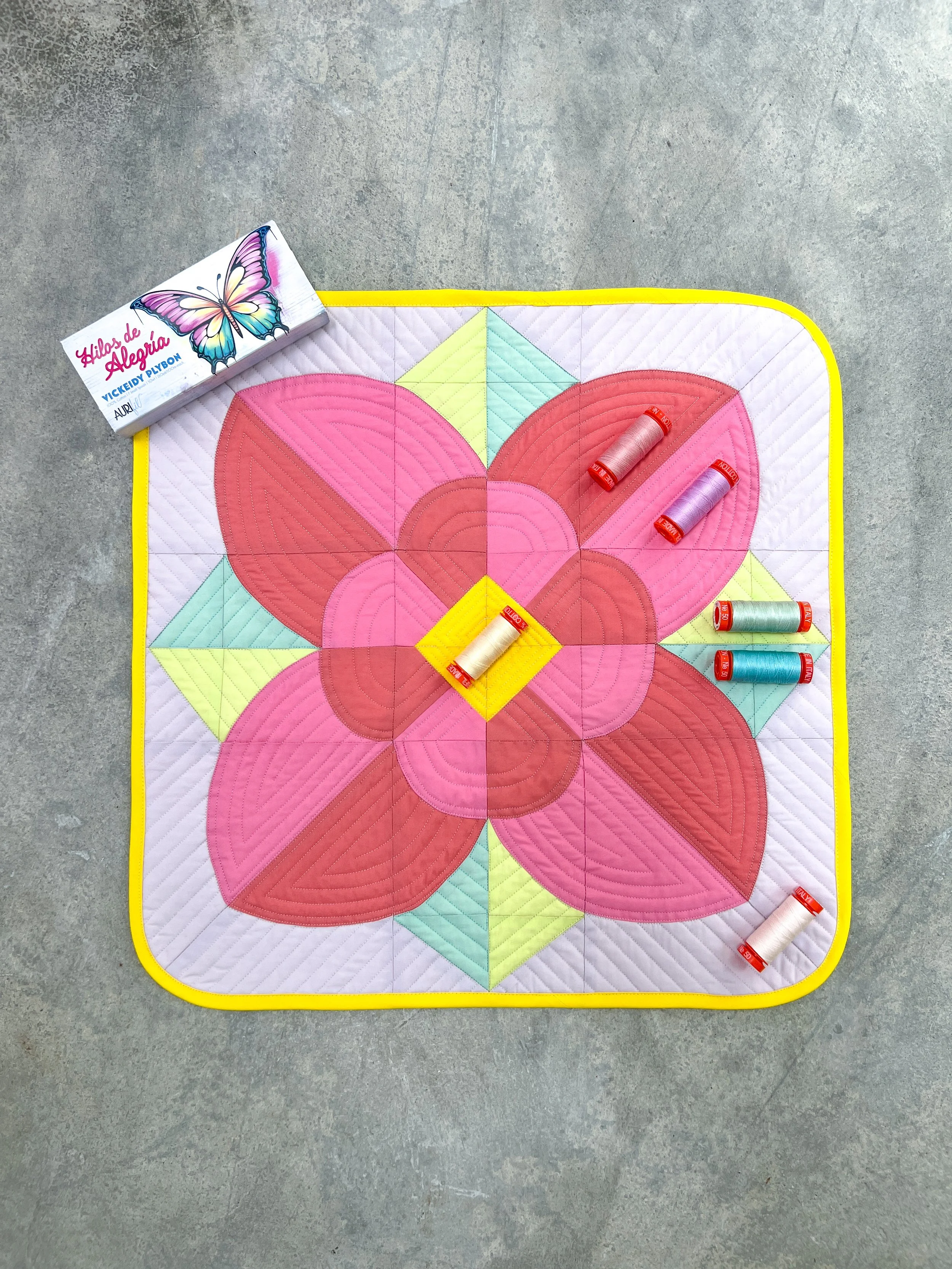 A quilt with a butterfly design in pink, yellow, and green colors, placed on a gray floor. Several spools of thread are on top of the quilt, and a box labeled 'Hilos de Alegria' with a butterfly illustration is nearby.
