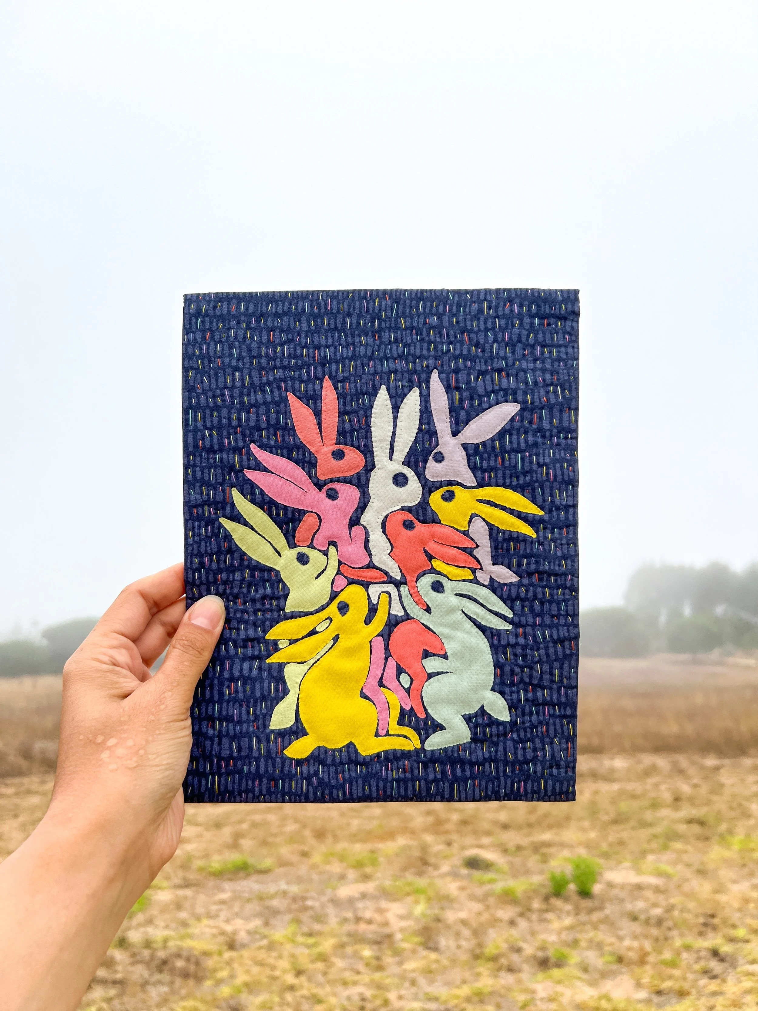 Hand holding a colorful fabric with a pattern of unicorns in pink, yellow, and white on a dark blue background.