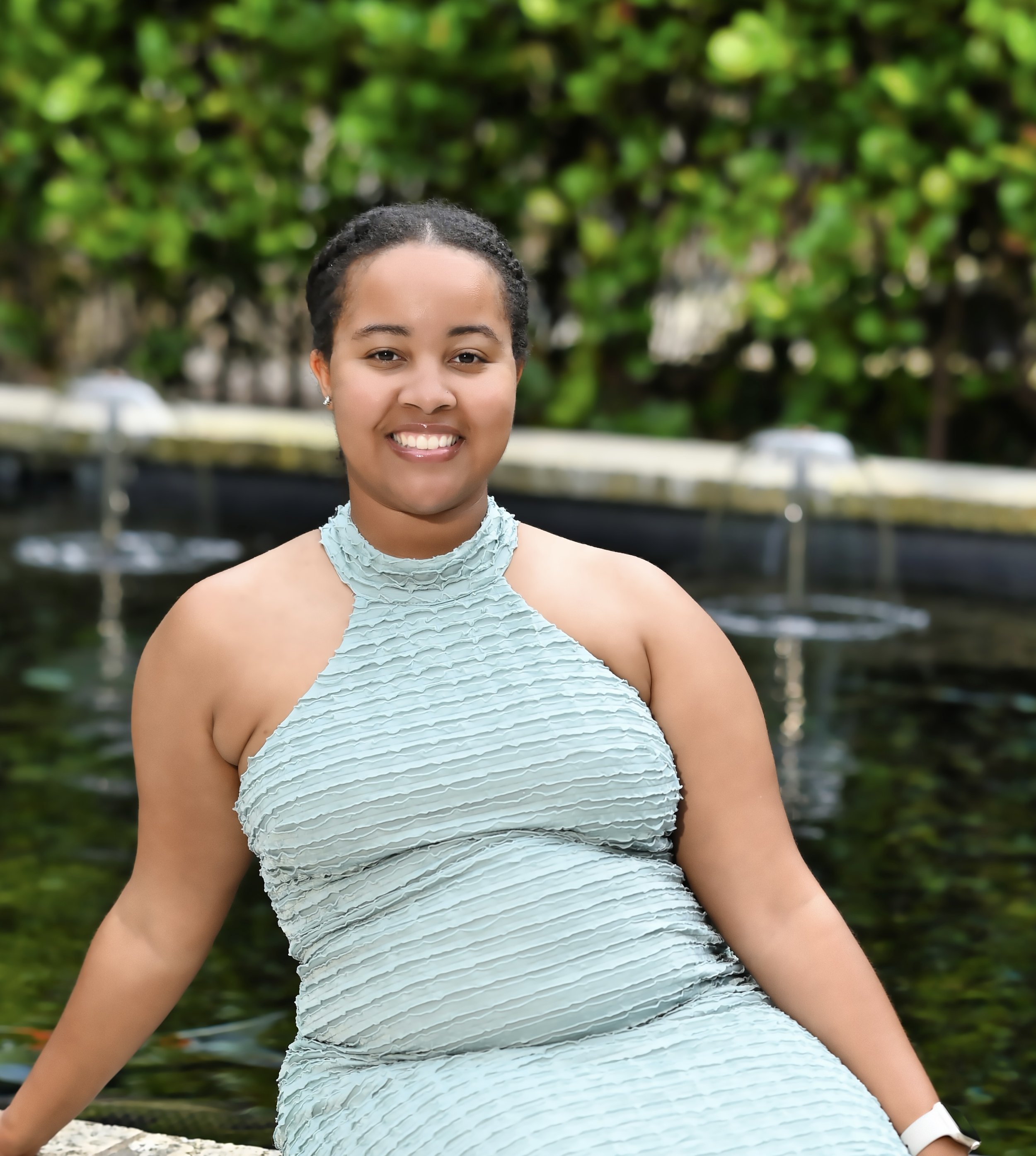 Close-up of a young woman smiling outdoors, wearing a light blue, textured sleeveless top, with a blurred background of green foliage and water.