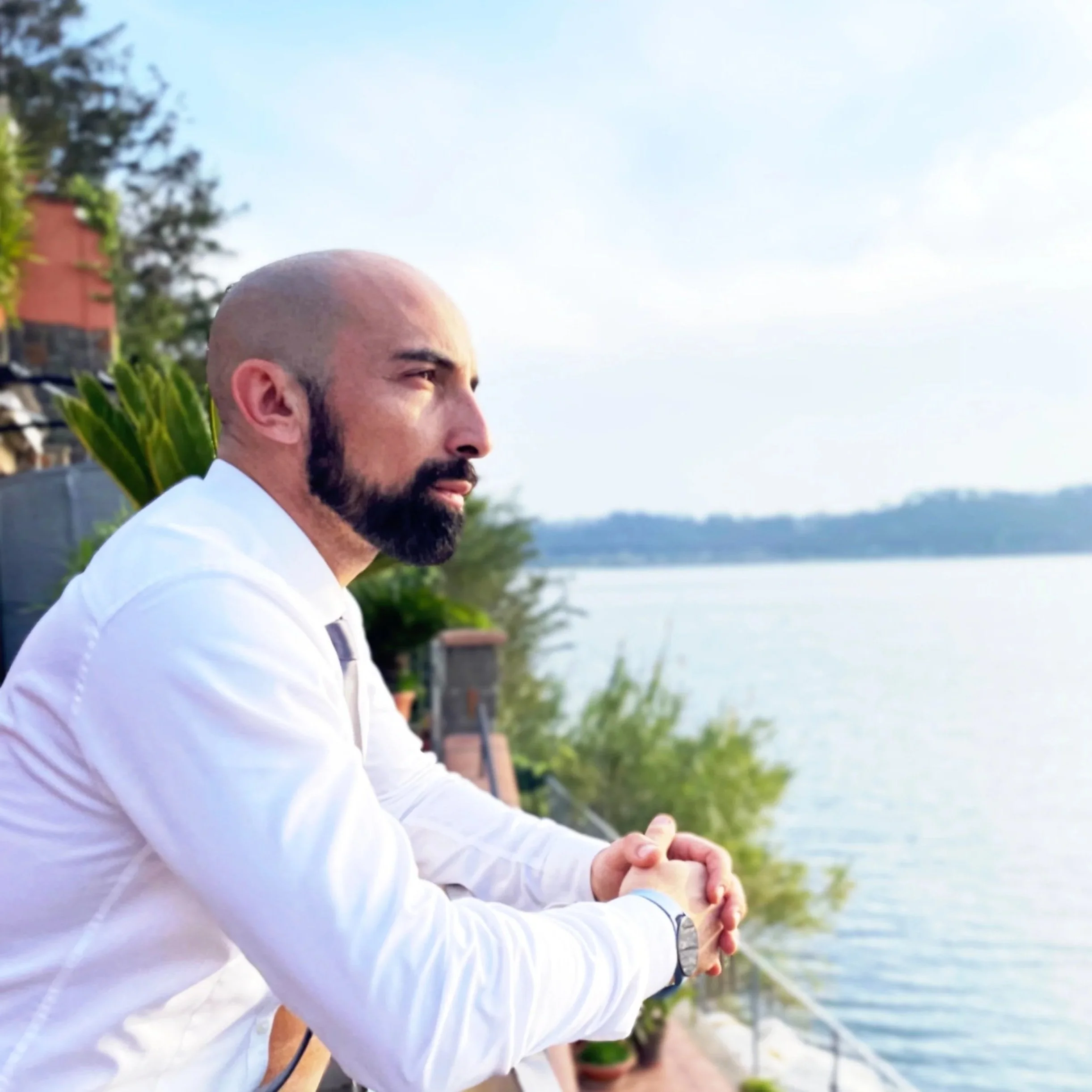 Un uomo con barba, vestito con camicia bianca, guarda pensieroso un lago dal parapetto, circondato da vegetazione e alberi, con un cielo sereno.