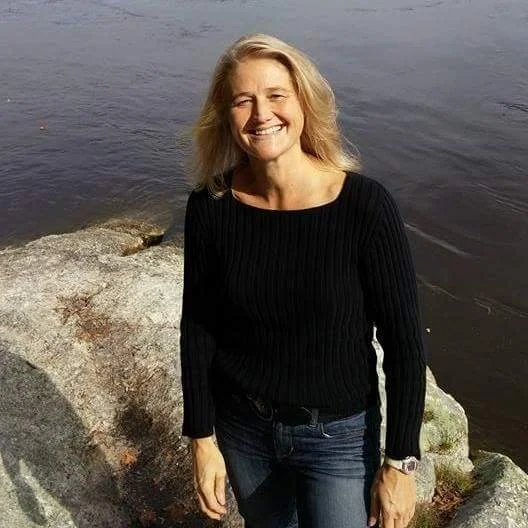 A smiling woman with blonde hair standing on rocks near a body of water, dressed in a black sweater and jeans.