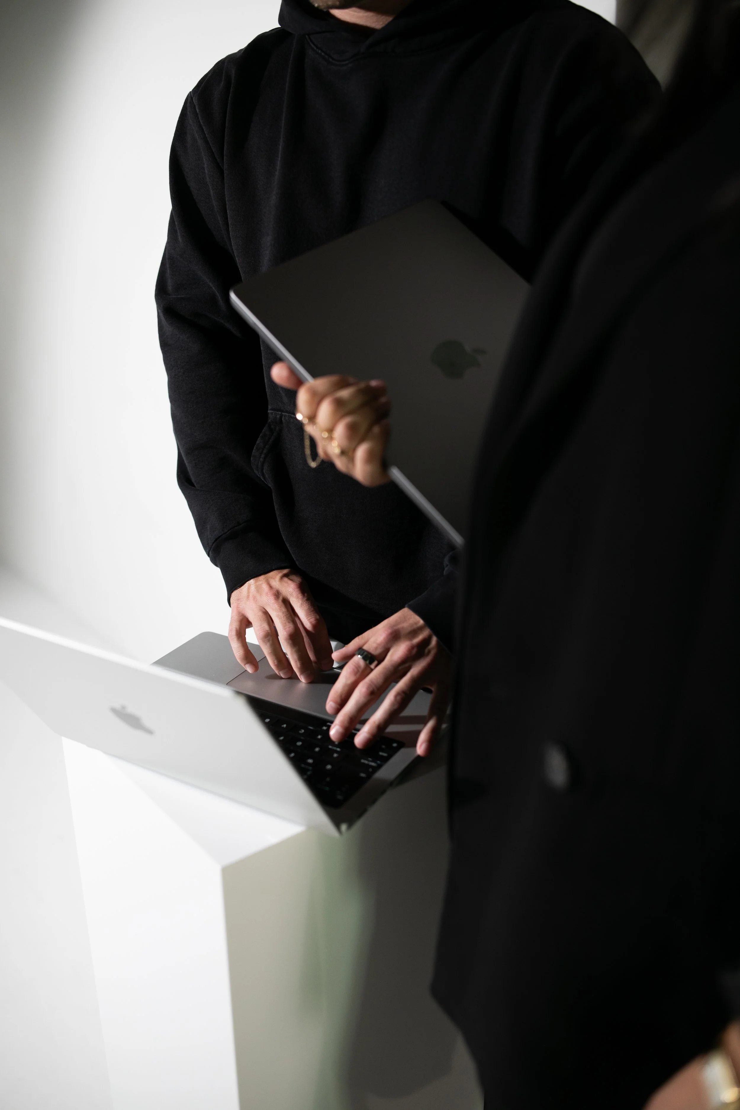 Person in black hoodie holding a closed laptop while standing at a white table with another open laptop, with a white wall in the background.